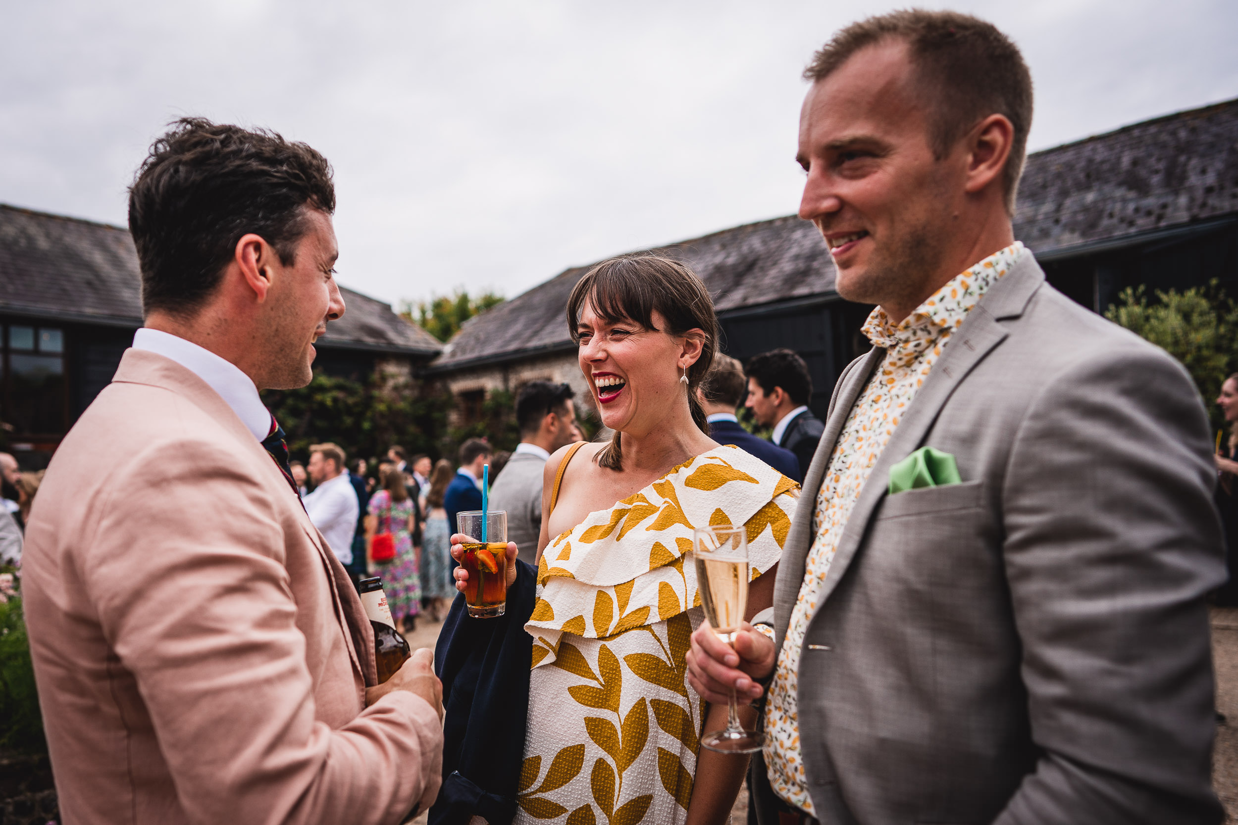 Three people in semi-formal attire chat and smile at an outdoor event, holding drinks.