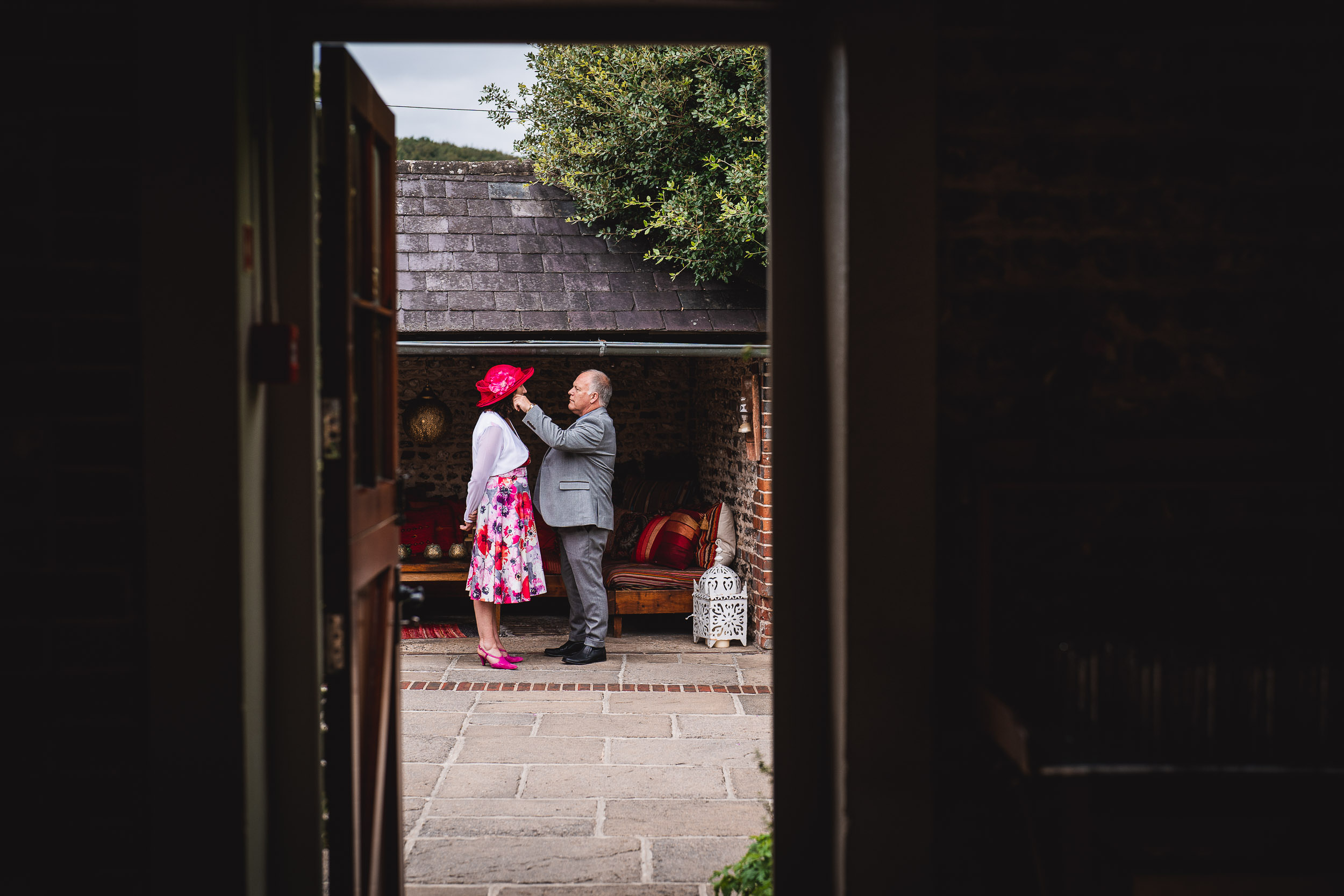 A man adjusts a woman's hat in a cozy outdoor setting, framed by a doorway. They stand on a stone patio with brick walls and greenery in the background.