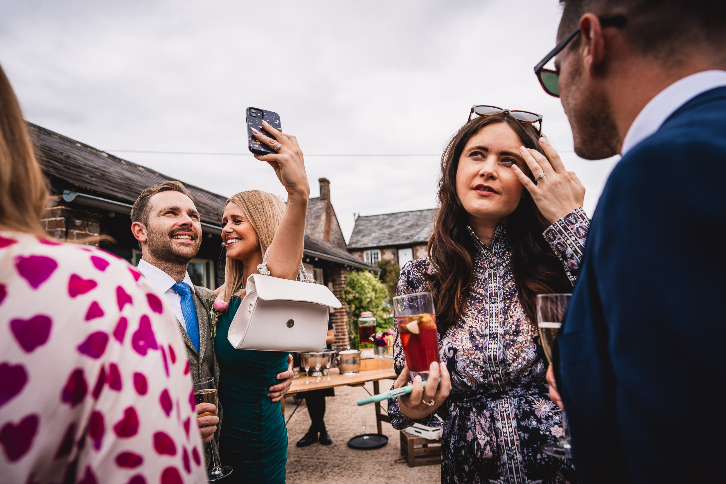 A group of people stand outdoors, some smiling for a selfie while others engage in conversation. One woman holds a drink.