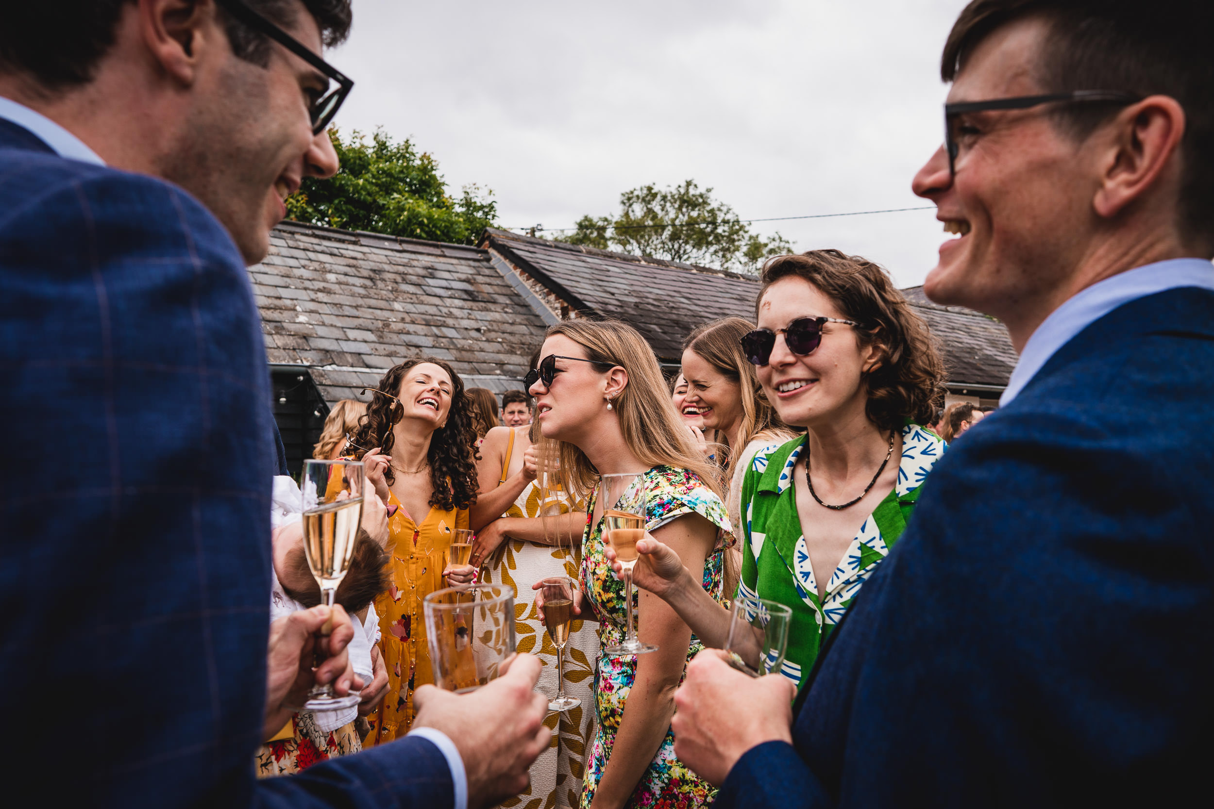 A group of people in formal attire are gathered outdoors, smiling and holding glasses. Some are wearing sunglasses. The scene appears lively and cheerful.