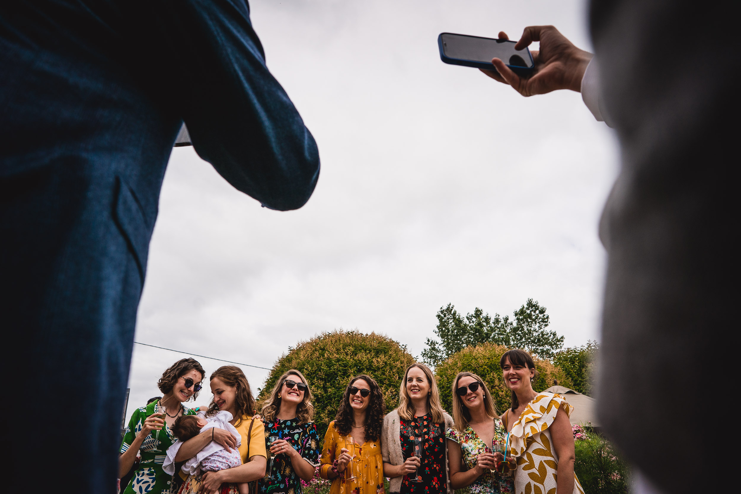 Group of people posing for a photo outdoors, with a person holding a baby and another taking a picture using a smartphone. Trees and cloudy sky in the background.