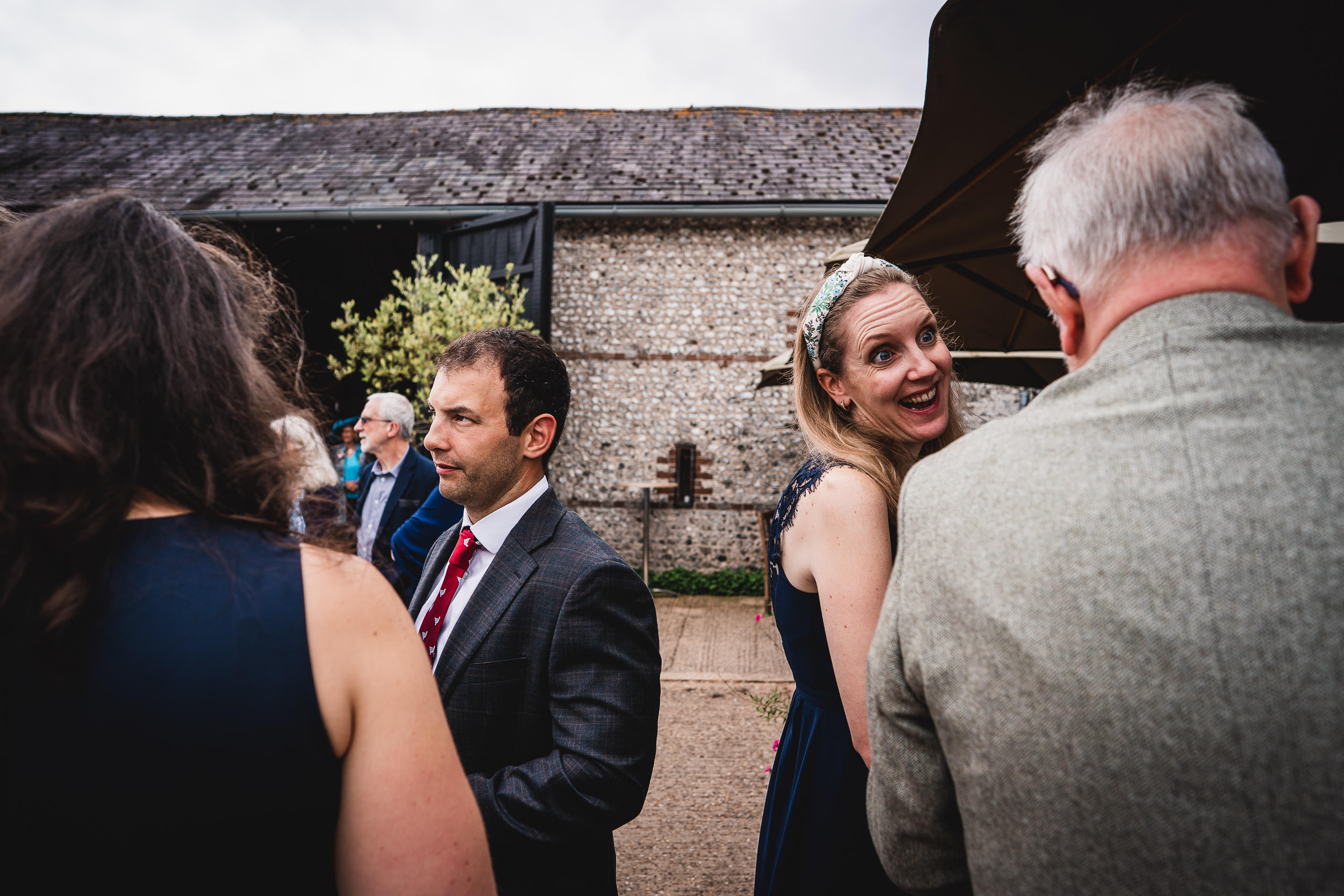 A group of people in formal attire stand outdoors near a stone building, engaged in conversation. One woman looks toward the camera, smiling.