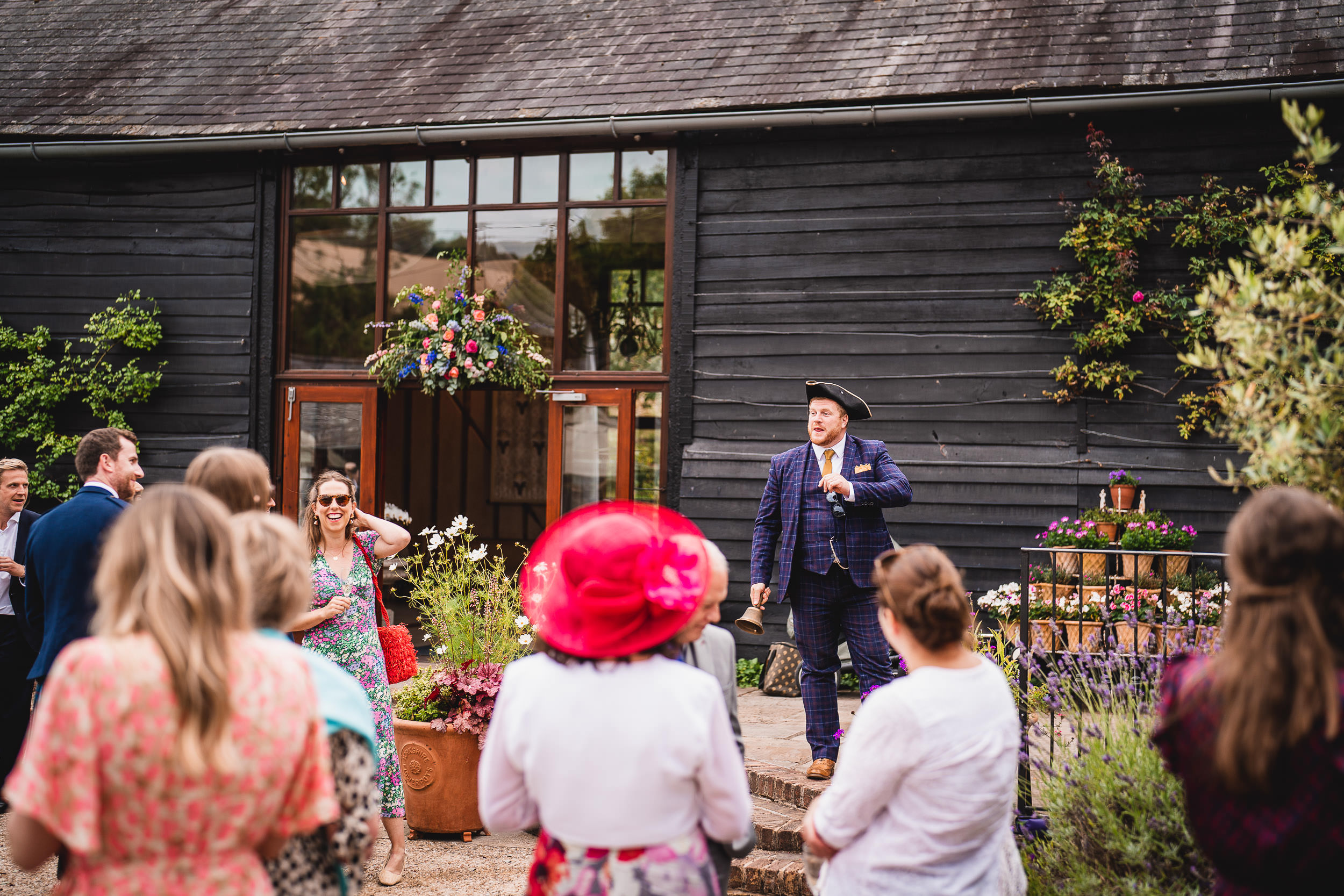 A man in formal wear gives a speech to a small crowd outside a rustic building with flowers and plants.