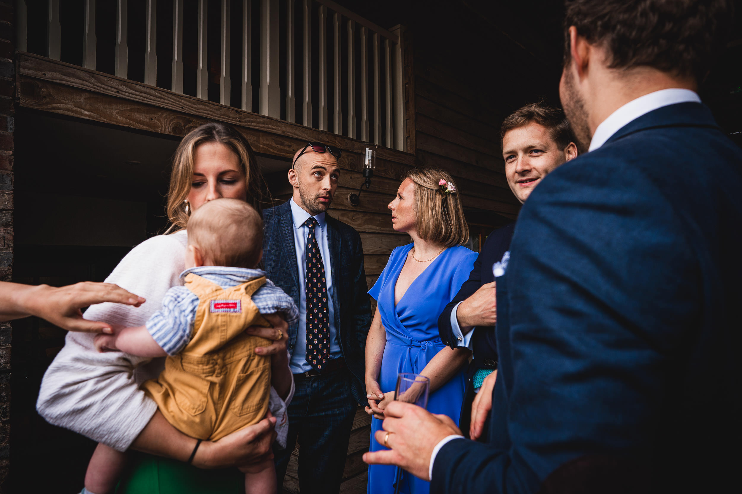 A small group of people in formal attire are gathered indoors, engaging in conversation. One person is holding a baby, and another is holding a drink.