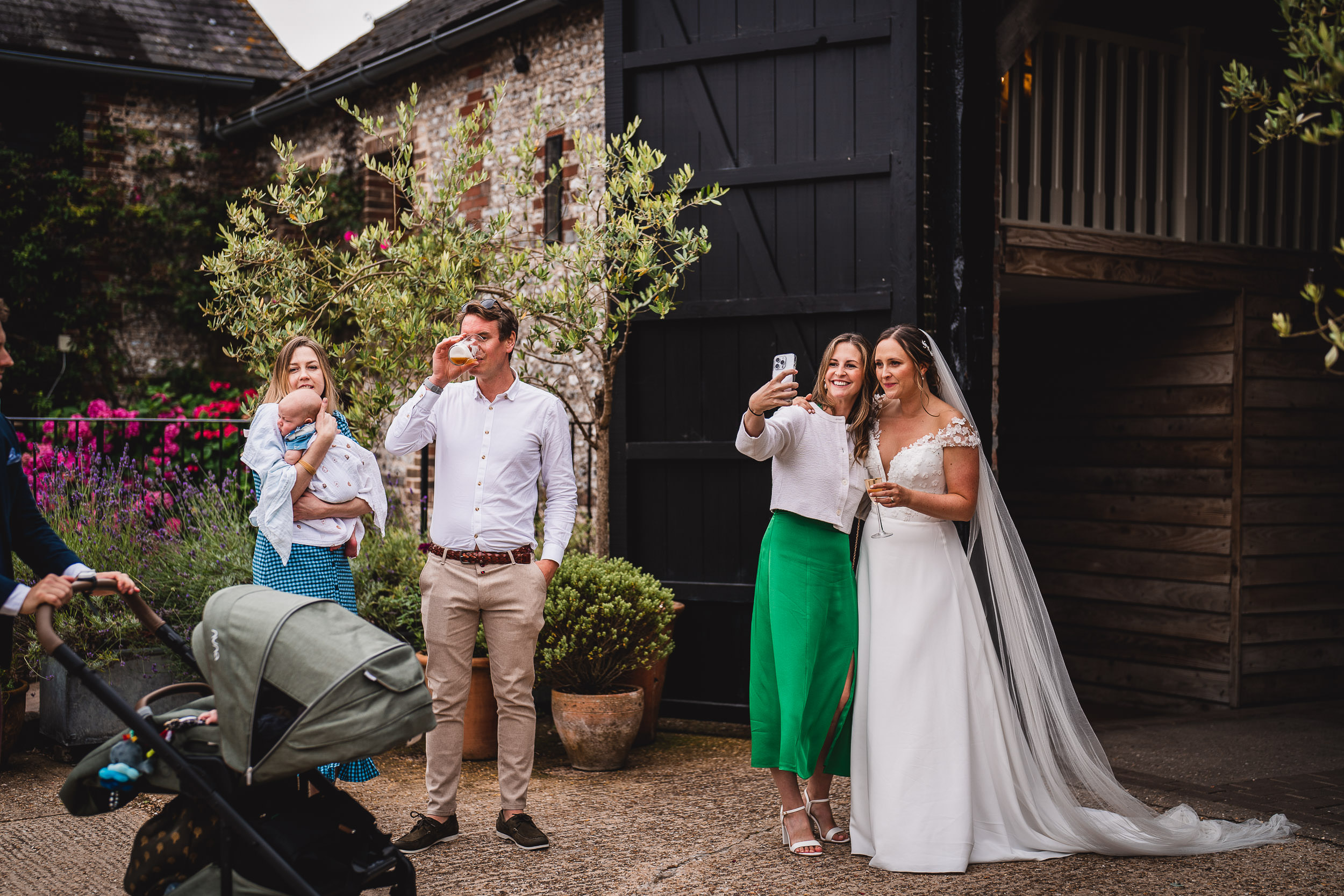 A group of people stand outdoors at a rustic venue. One person holds a baby, while two others take a selfie. A stroller is in the foreground. The bride wears a white dress and veil.