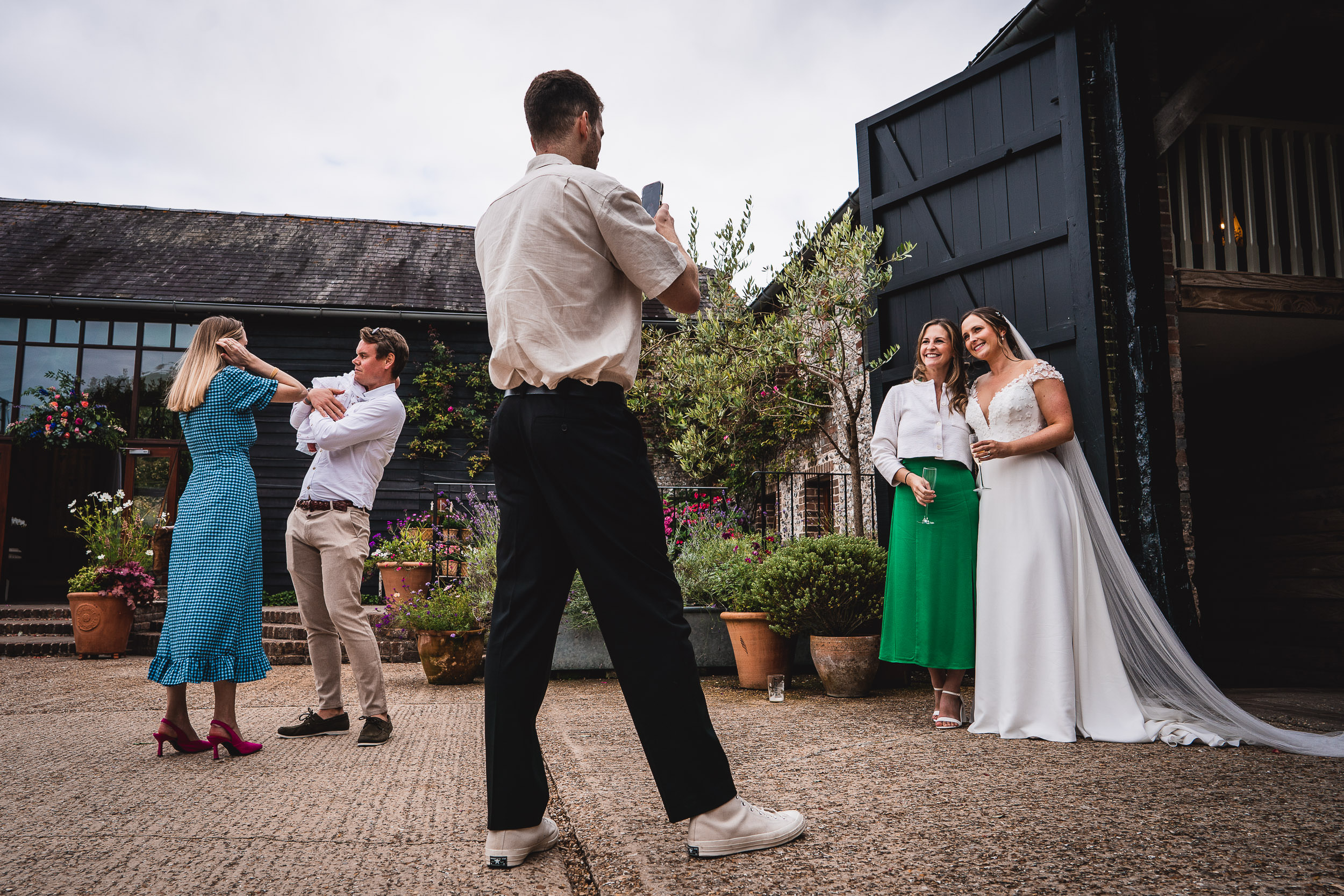 A person photographs two women in wedding attire while another couple poses playfully outside a barn-style venue.