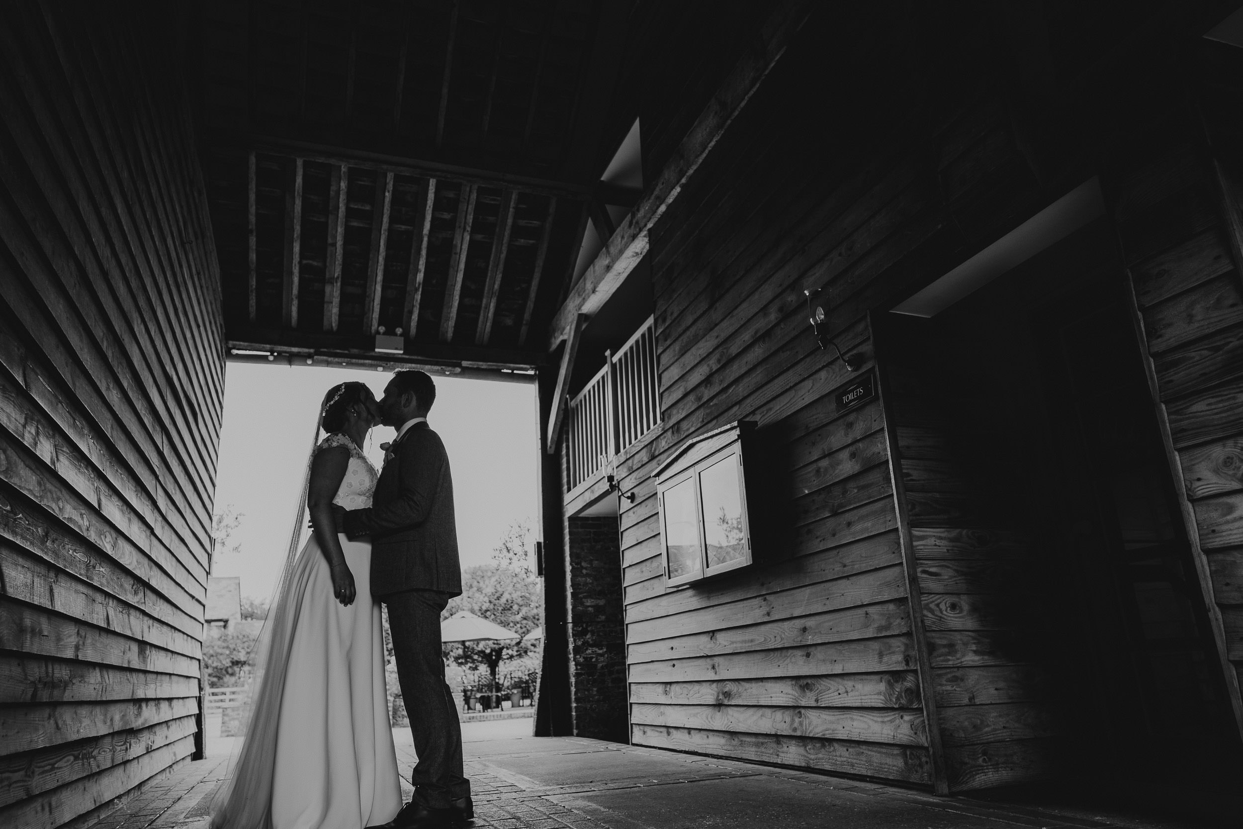 A bride and groom stand facing each other under a wooden archway, silhouetted against the light.