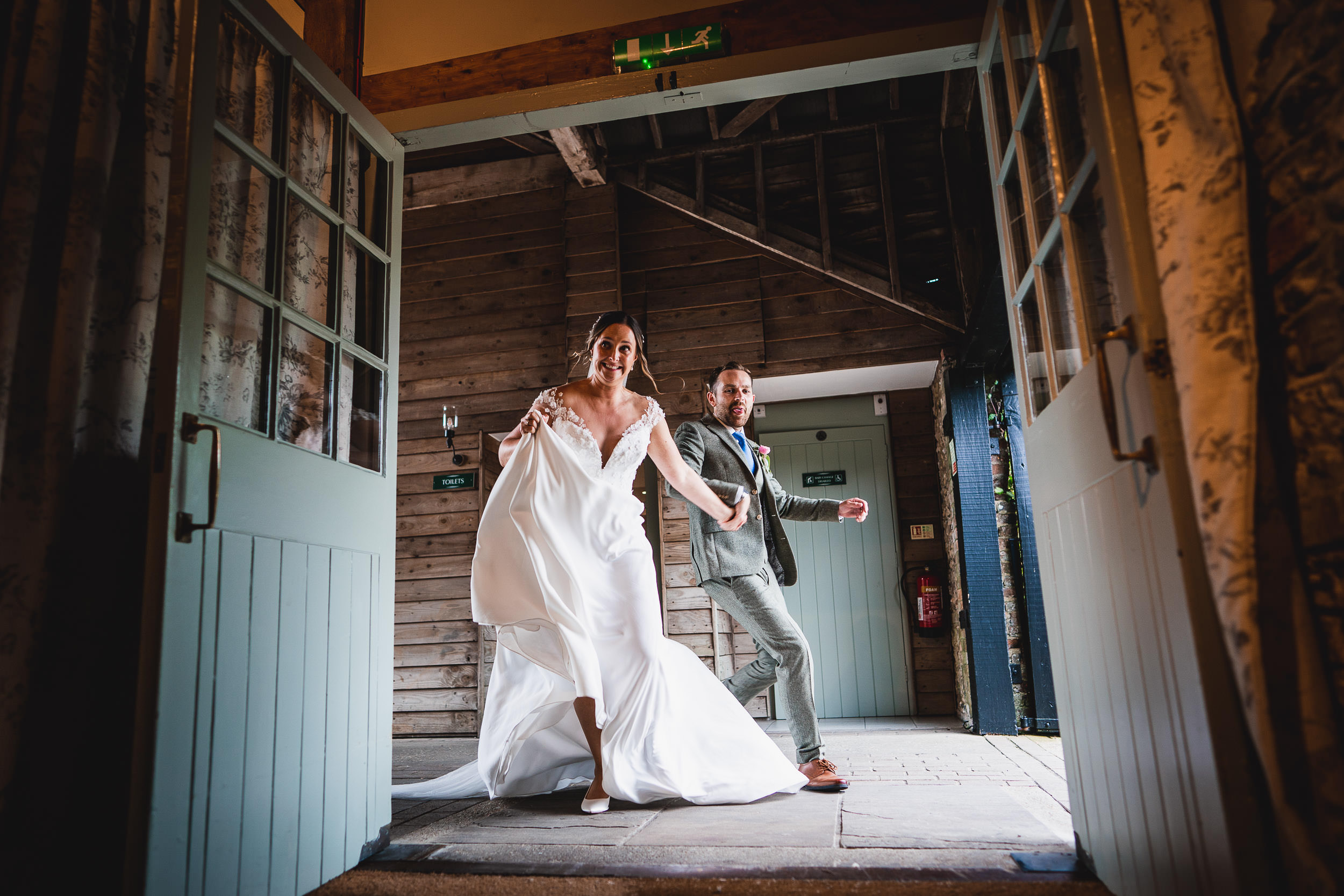 A couple joyfully enters a rustic venue. The woman wears a flowing white dress, and the man is in a grey suit. They appear to be in a celebratory mood.