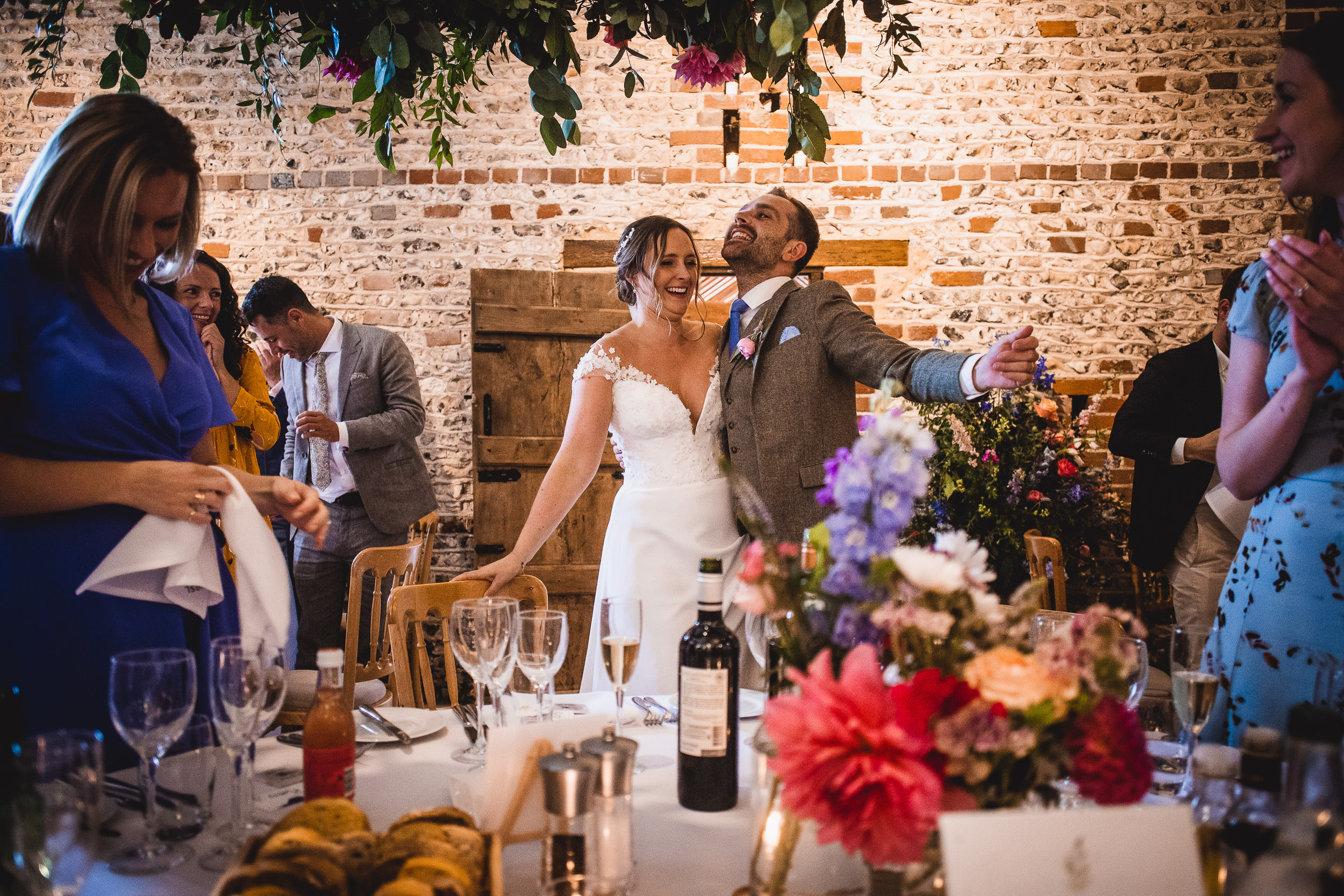 Bride and groom smiling joyfully at a wedding reception, surrounded by guests and a decorated table with flowers and drinks.