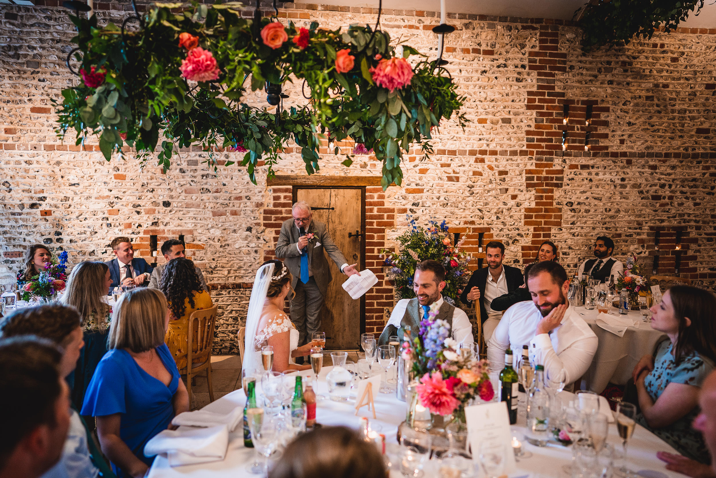 A speaker addresses a seated audience at a wedding reception. The venue features brick walls and floral decorations. Guests listen attentively.