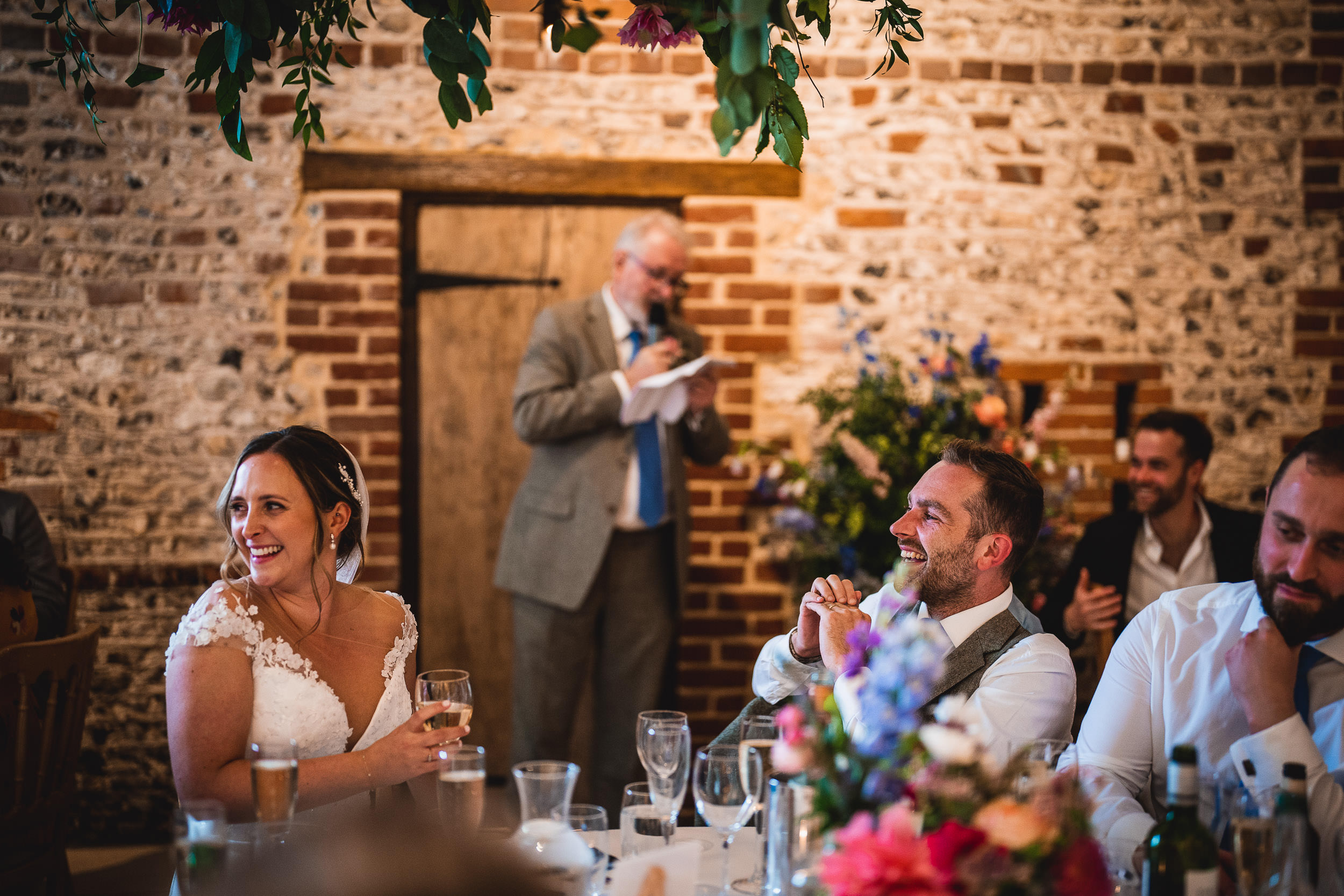 A bride and groom sit at a table, smiling and listening to a man speaking in a suit. The setting has rustic brick walls and floral decorations.