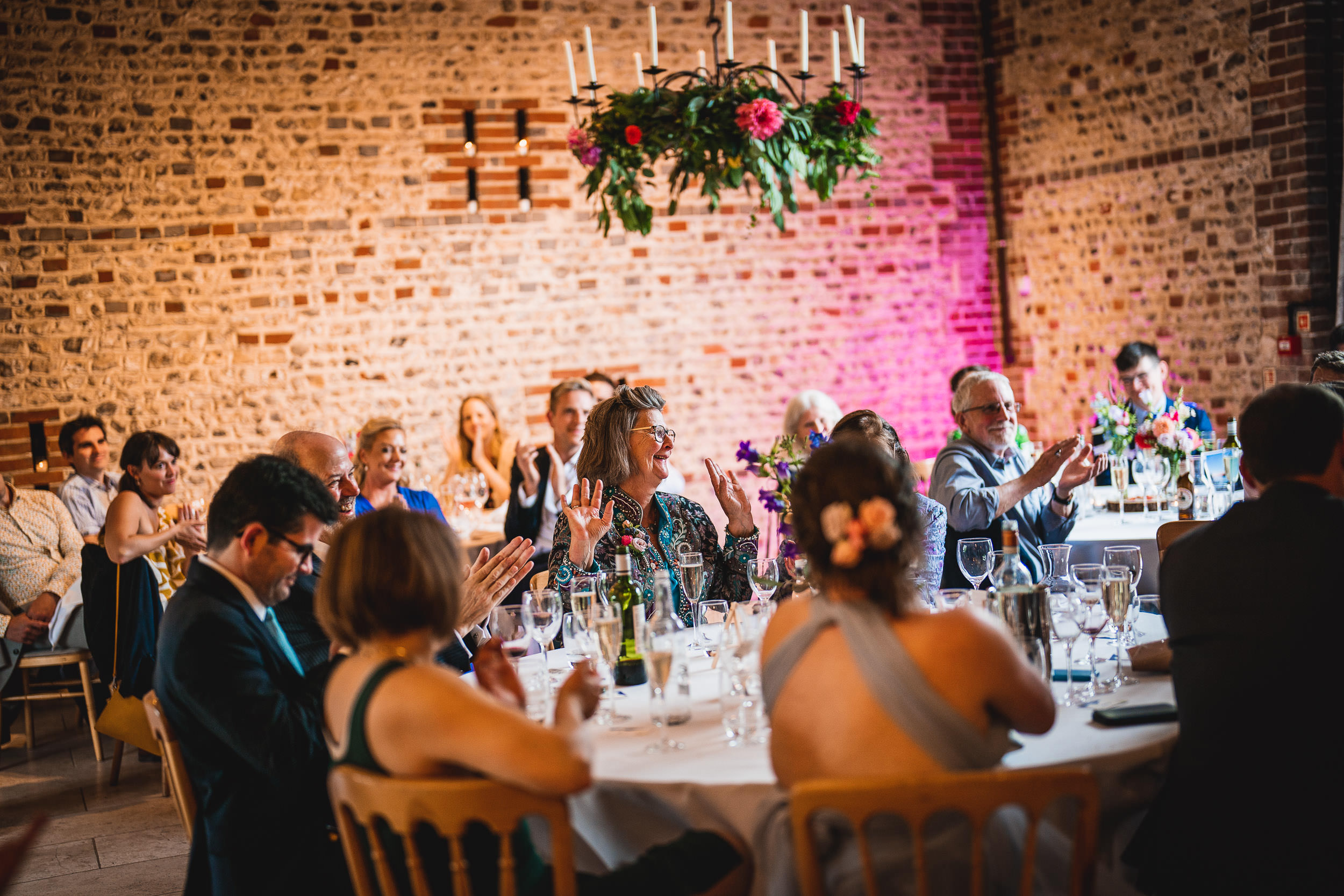 Guests seated at round tables in a rustic venue clap and watch a presentation. A brick wall and chandelier with flowers are in the background.