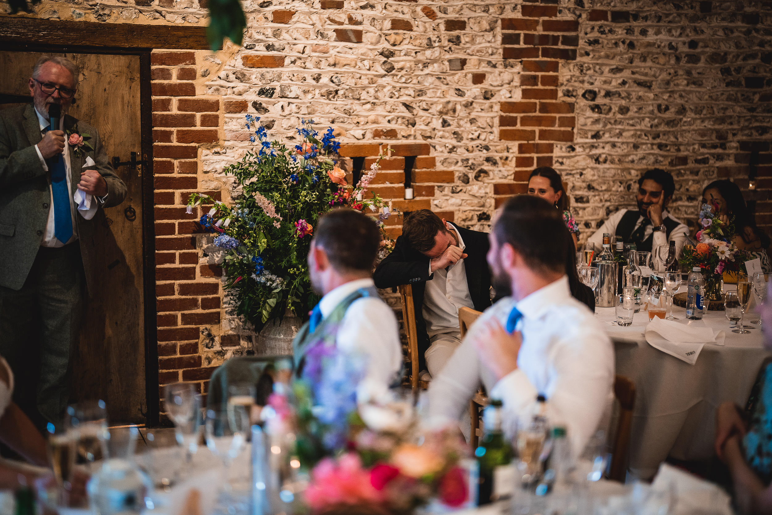 A wedding reception with guests seated at tables, a man giving a speech, and another person appearing emotional or overwhelmed. Tables have flowers and drinks.