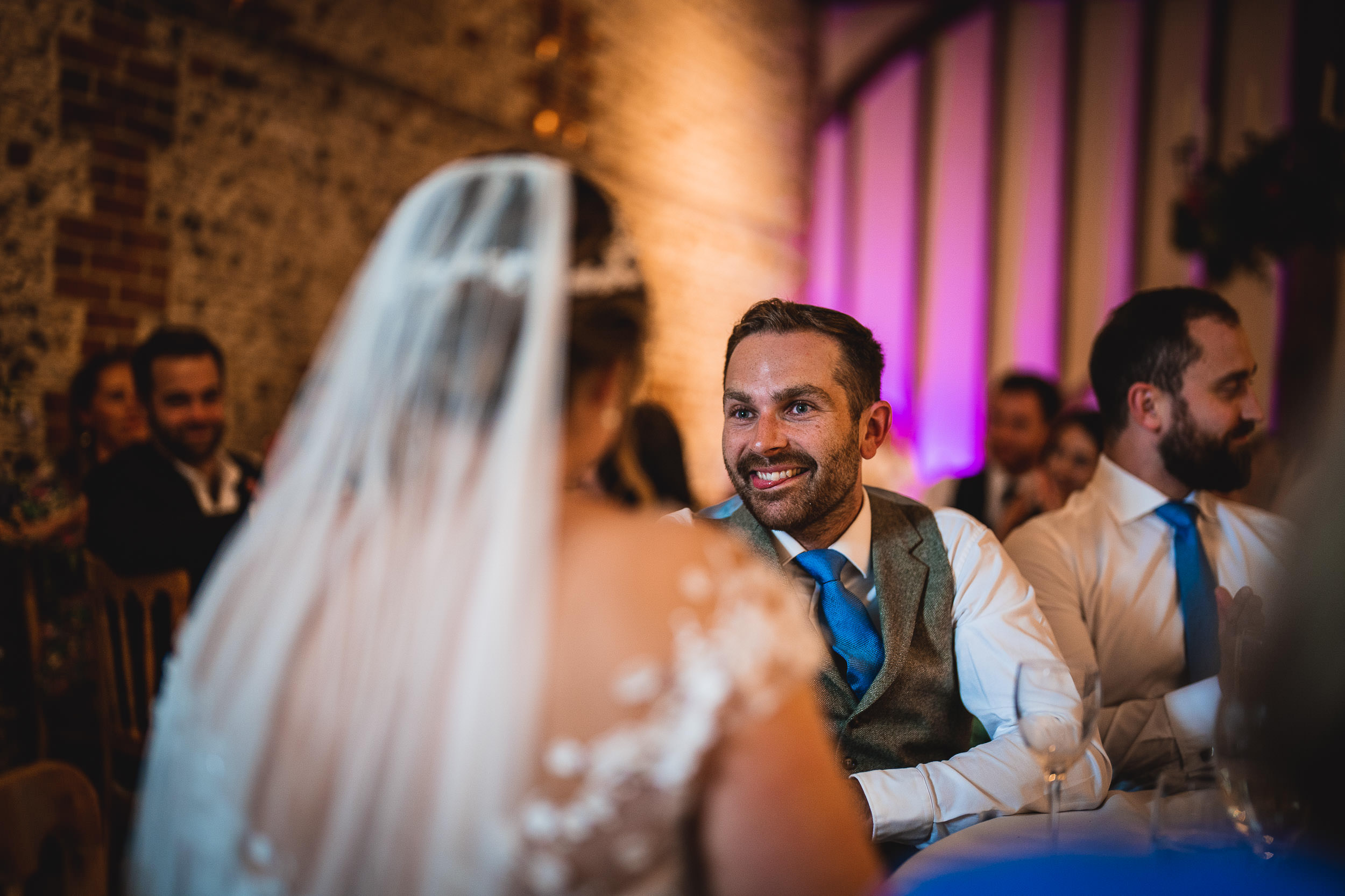 A man in a suit, sitting at a table, smiles at a woman wearing a wedding dress and veil.