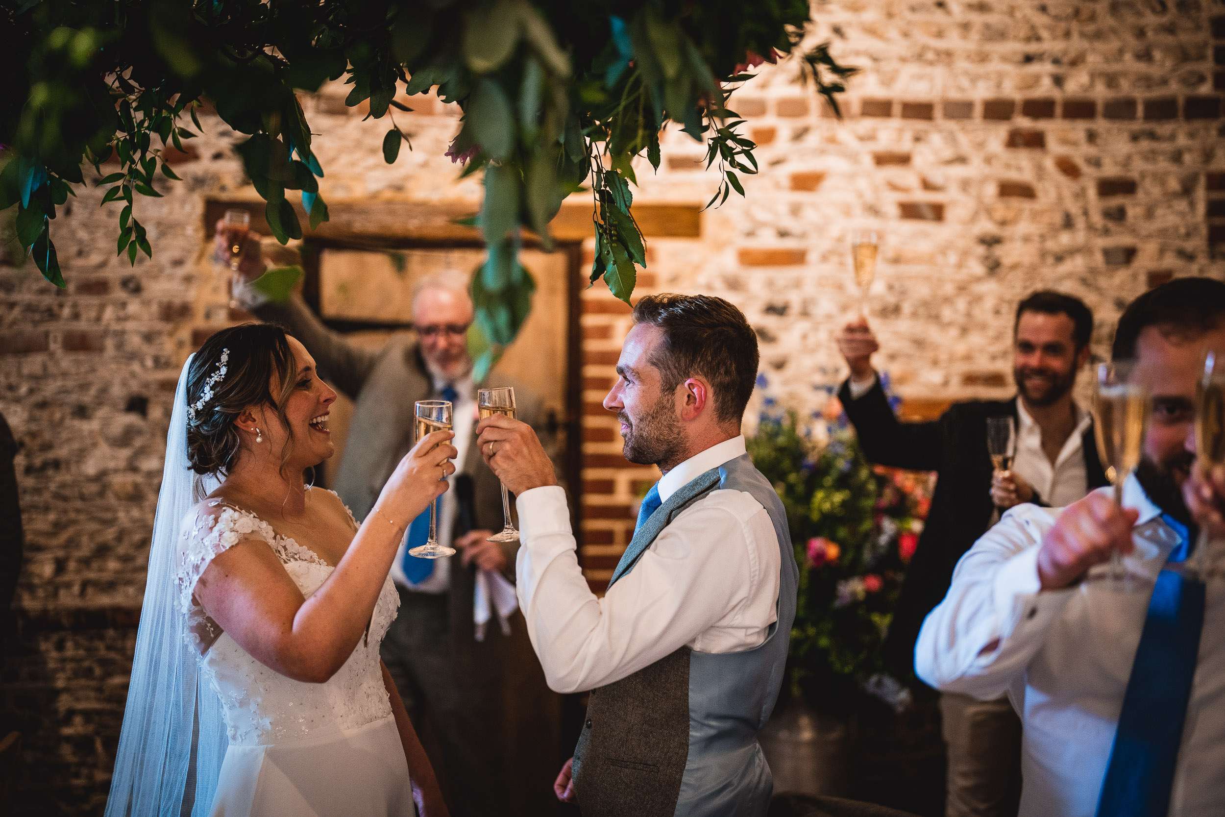 Bride and groom smiling and toasting with champagne glasses at a wedding reception, surrounded by guests.