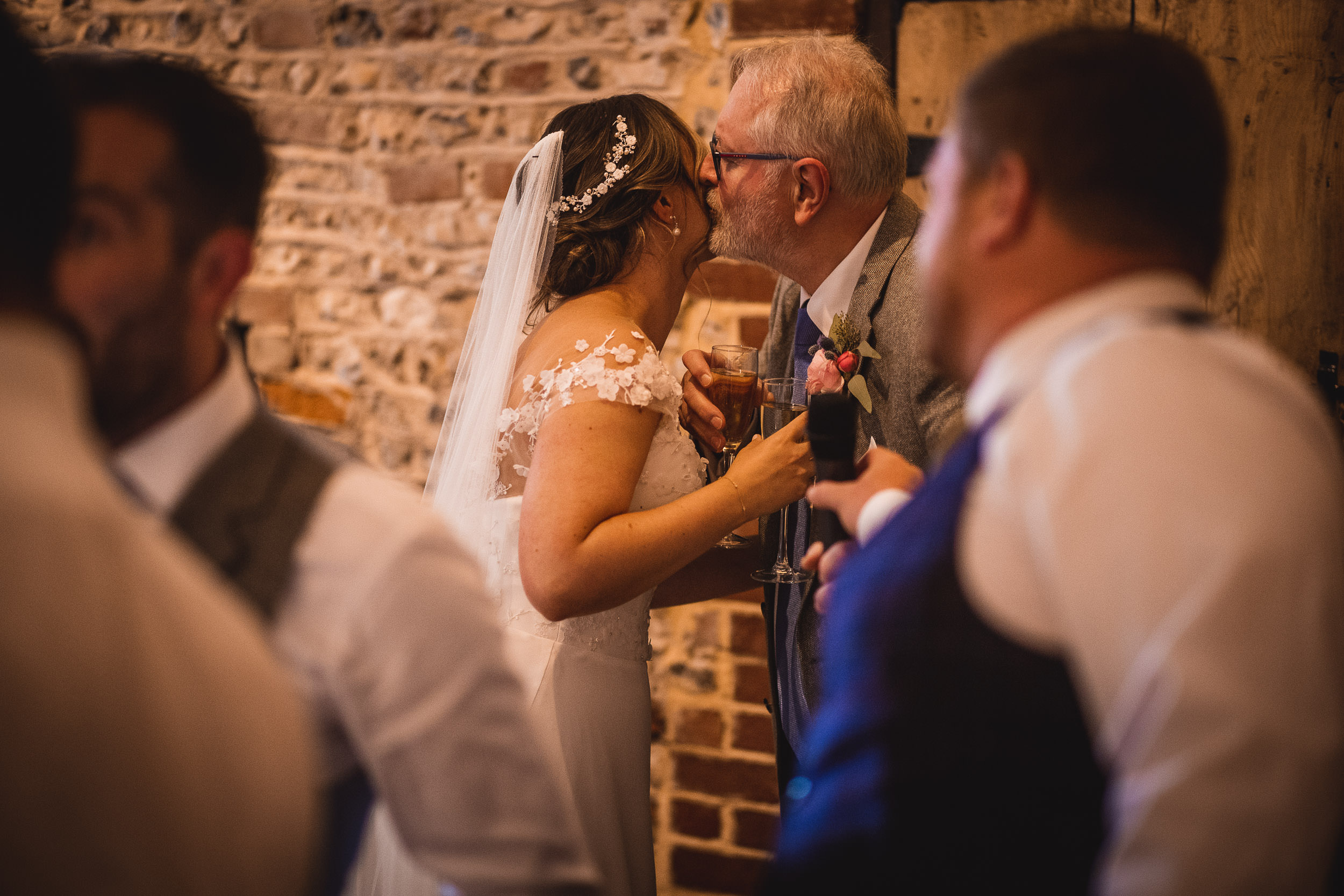 A bride and an older gentleman share a kiss at a wedding reception, both holding drinks. Other guests are seen in the foreground.