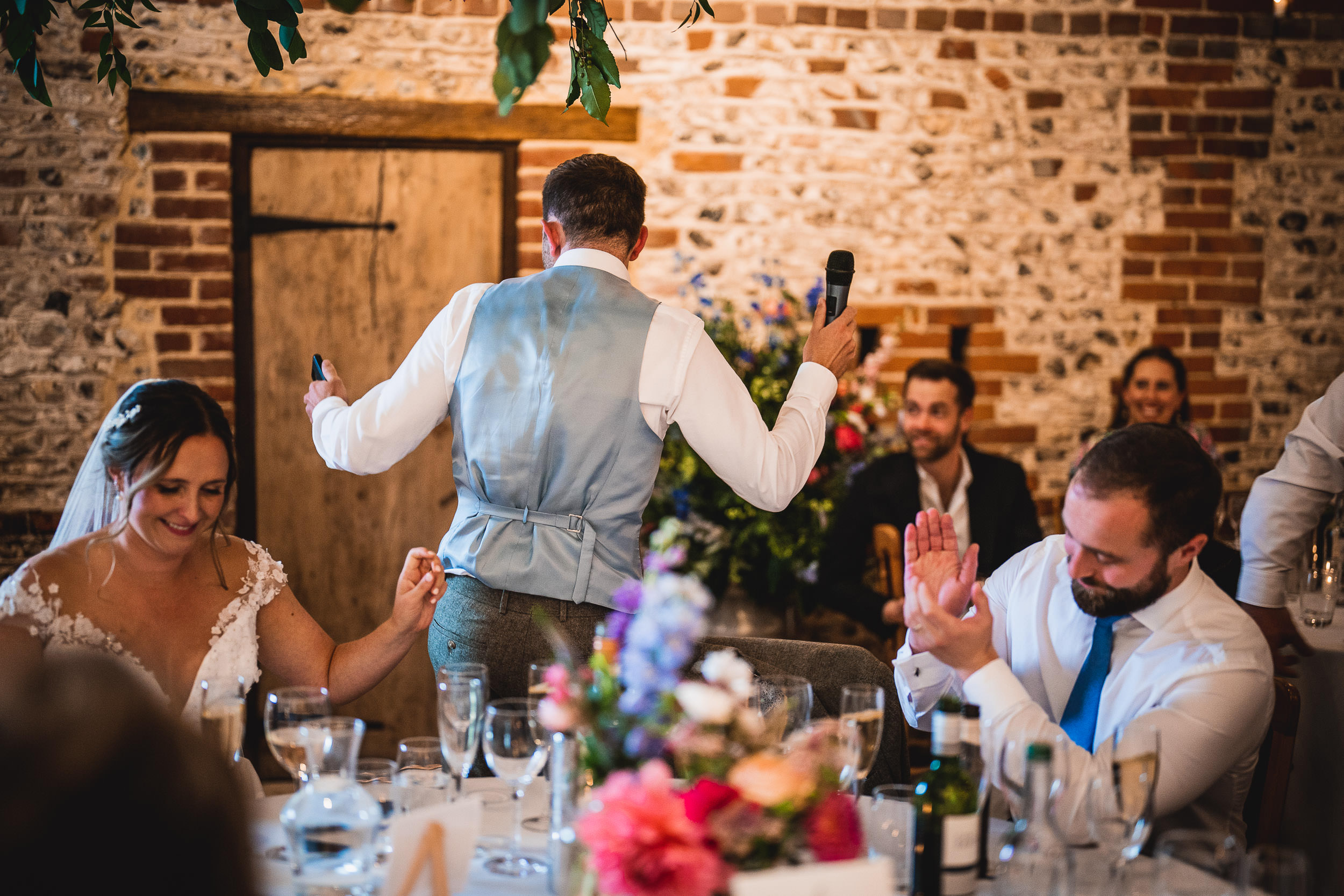 A groom in a blue vest speaks to wedding guests, holding a microphone. A bride sits smiling while a guest claps nearby. The setting features brick walls and floral arrangements.