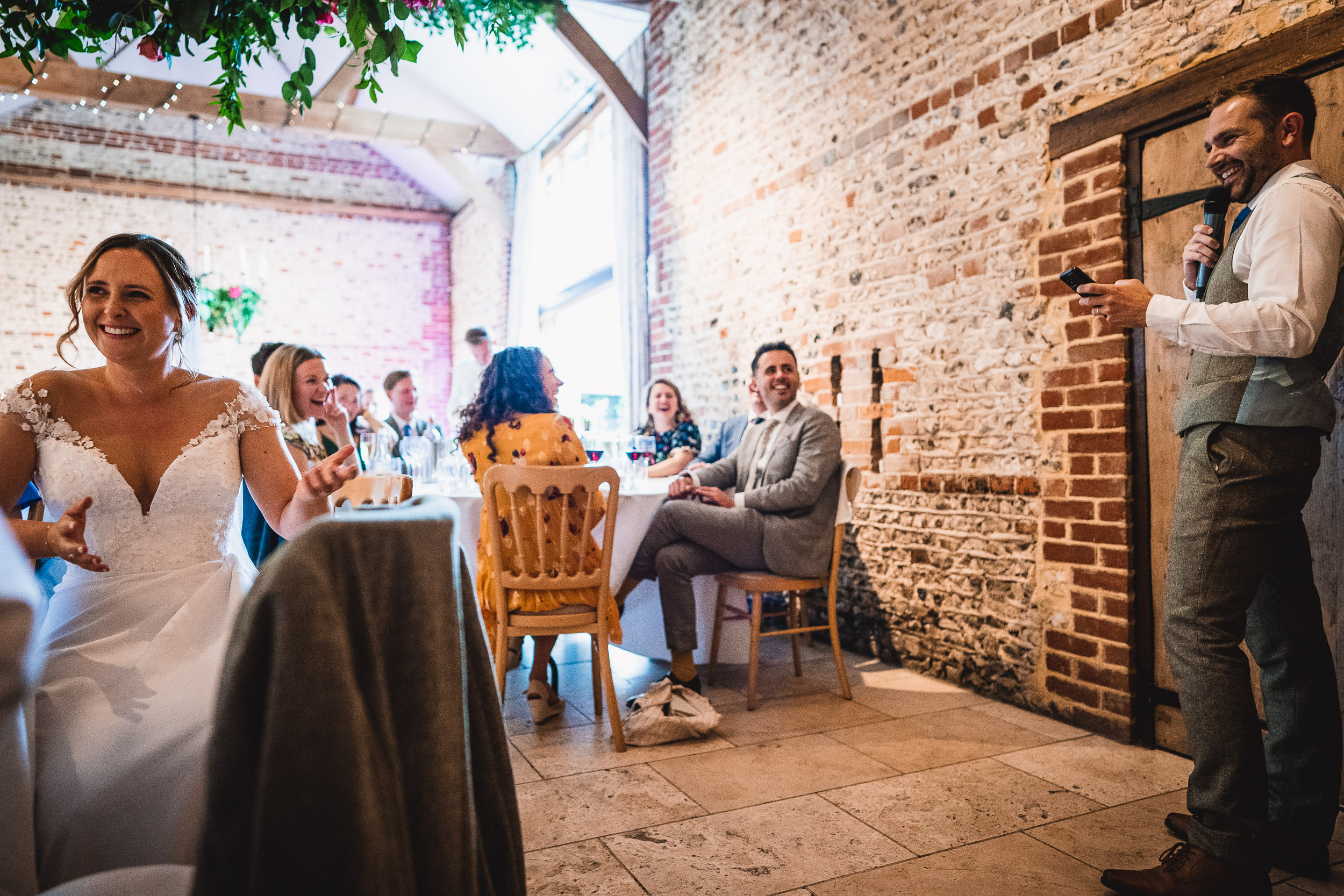 A bride and groom are seated in a rustic hall, while a man in a vest delivers a speech using a microphone. Guests enjoy the event. Decor includes greenery and exposed brick walls.