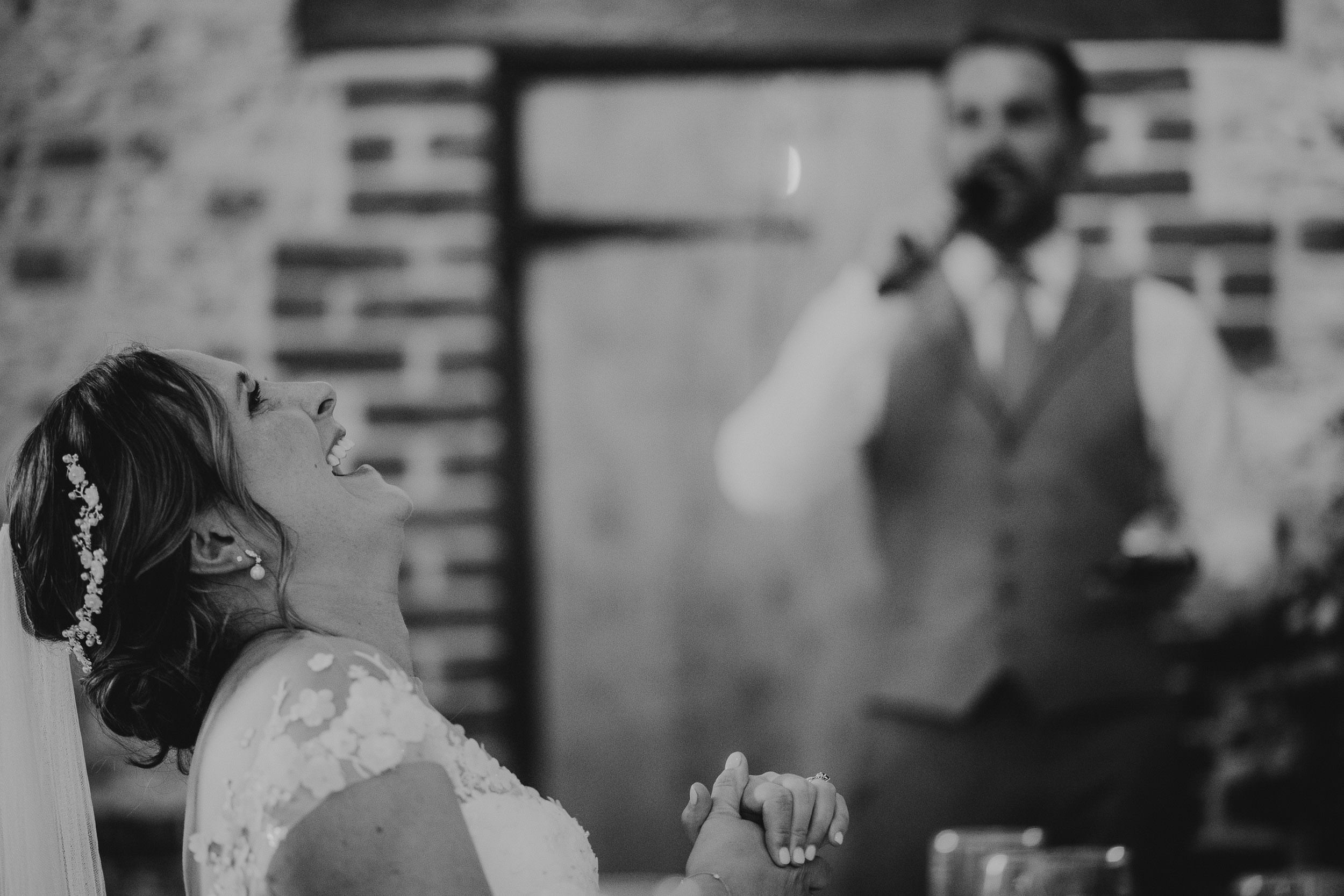 A bride laughs joyfully while a man in a vest speaks into a microphone during a wedding reception.