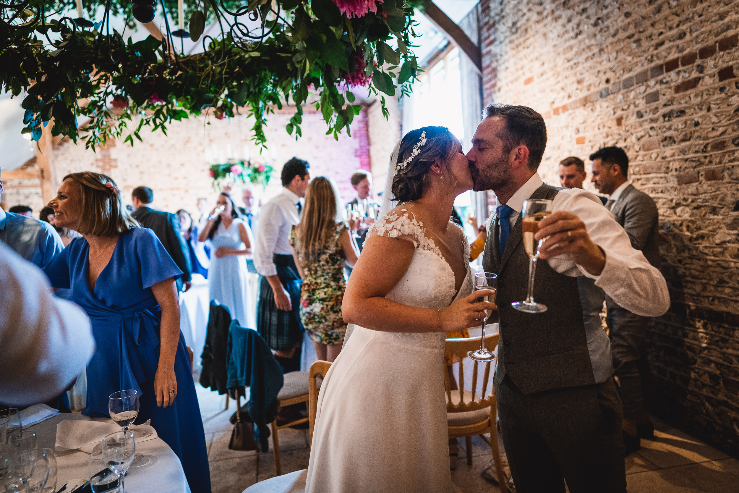 A bride and groom kiss while holding champagne glasses at a wedding reception, surrounded by guests in a decorated venue.