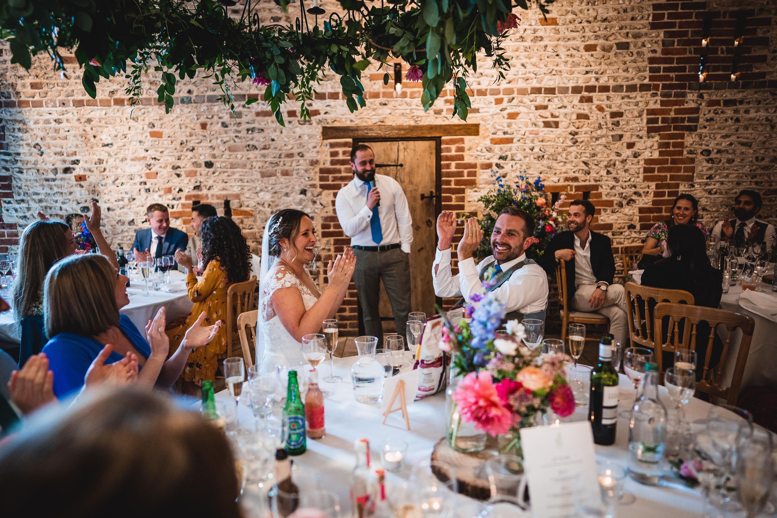 A wedding reception with a bride and groom sitting at a table, surrounded by guests. A man is standing and speaking, possibly giving a speech. The brick wall and flowers decorate the setting.