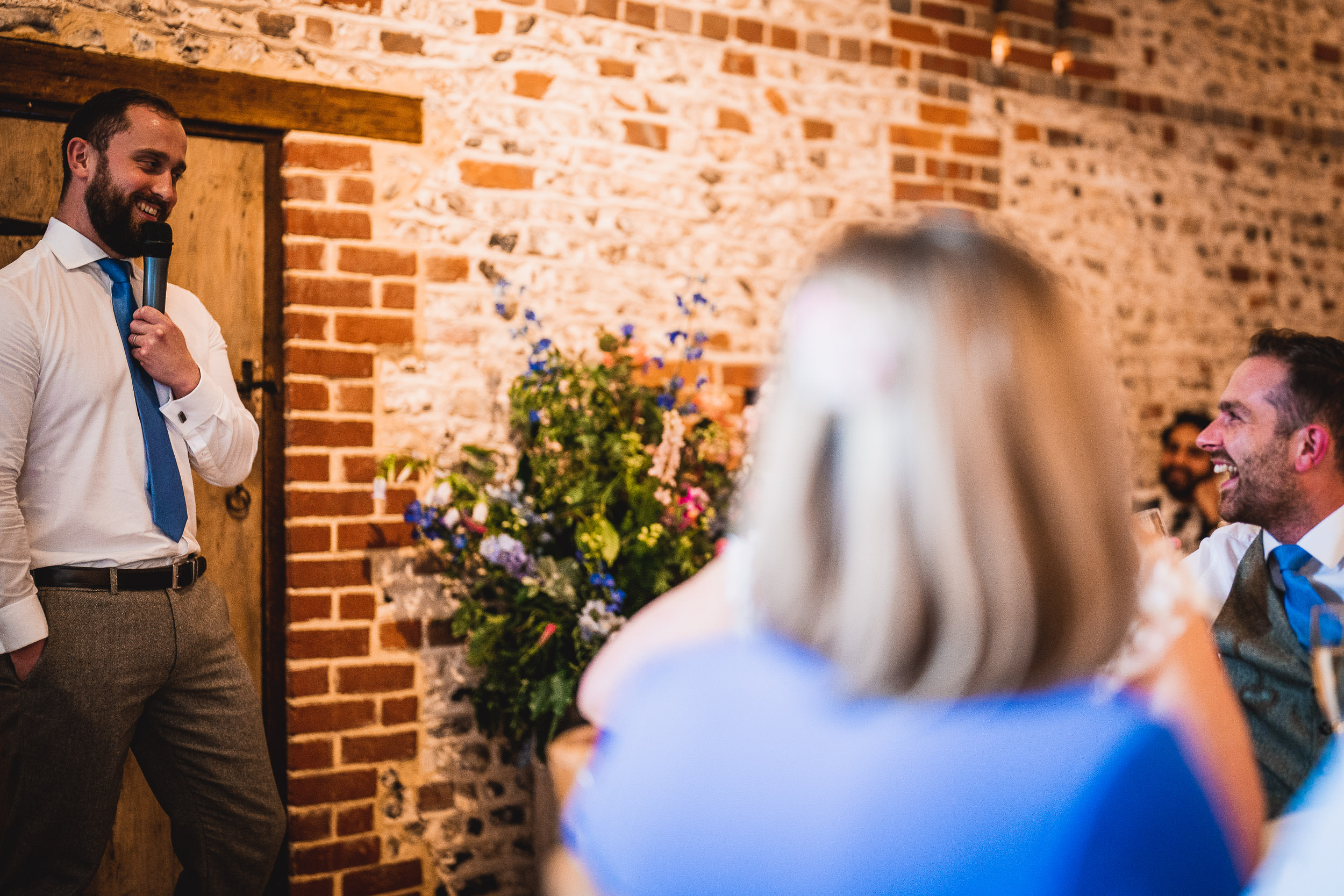 A man stands holding a microphone, smiling, while speaking to a seated group at a gathering in a rustic brick venue with floral decorations.