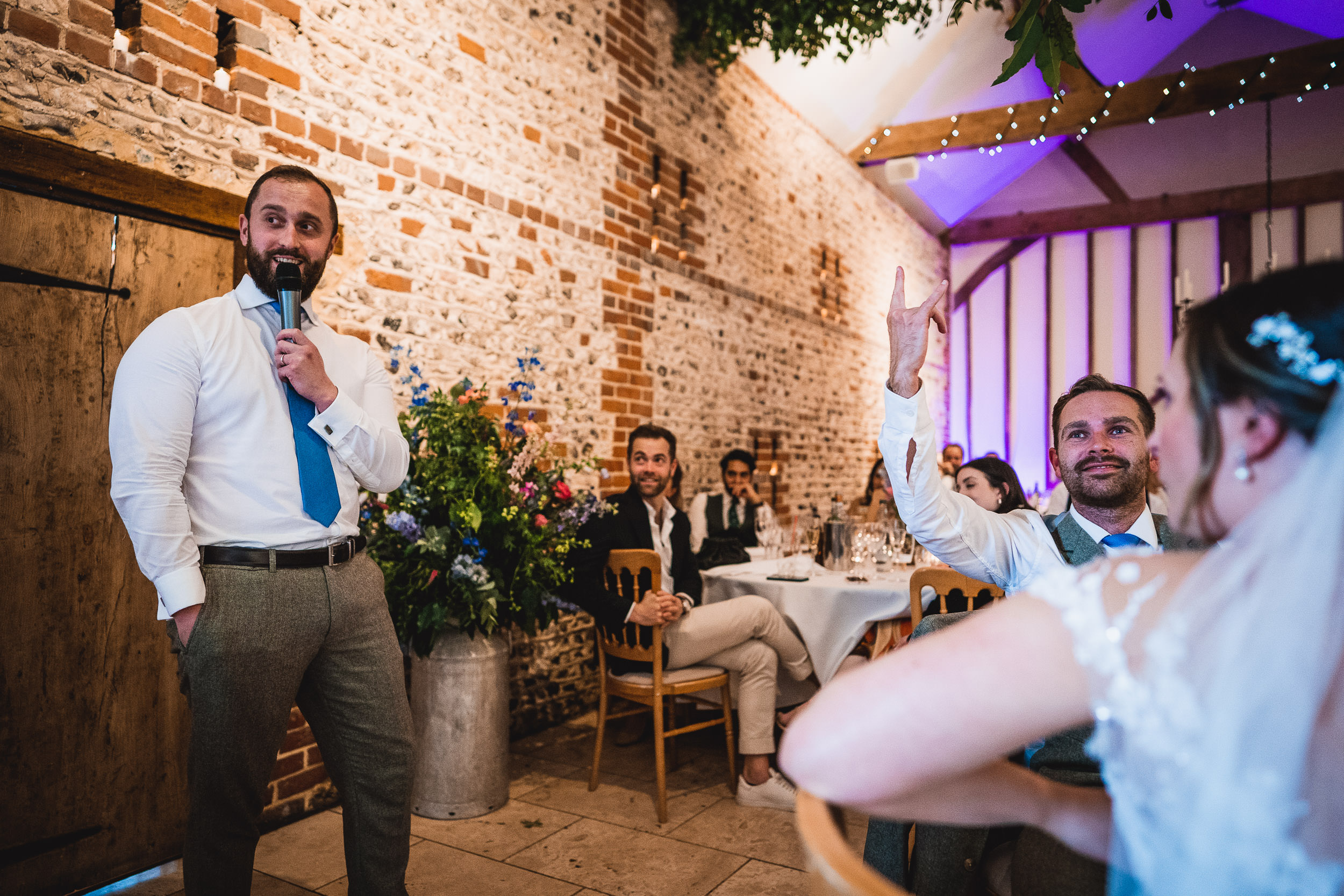 A man in a white shirt and blue tie is speaking into a microphone at an event. Another man raises his hand while seated near a person in a wedding dress. The venue has exposed brick walls.