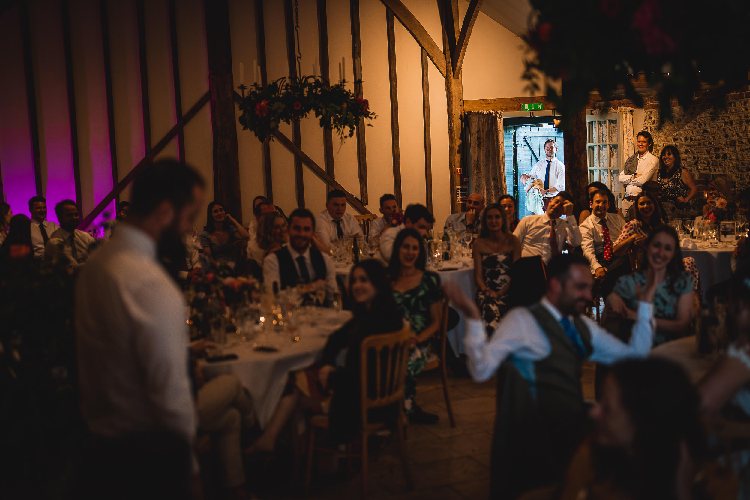 A man stands in a dimly lit room in front of seated guests, with wooden beams and floral decor visible.