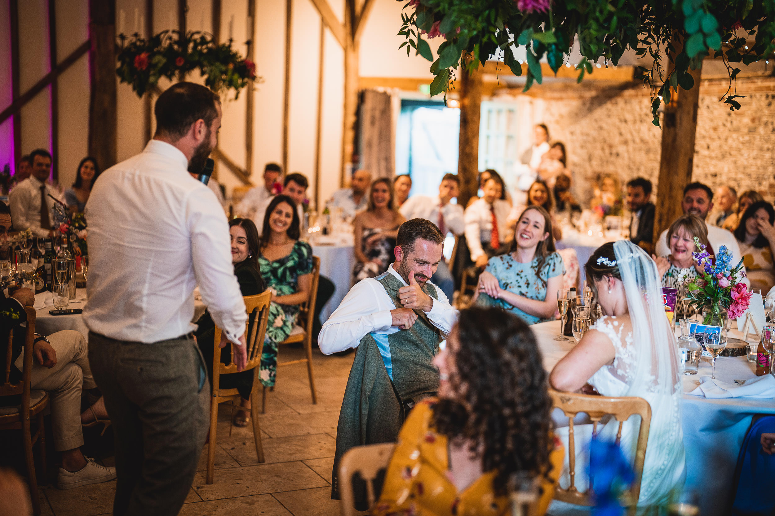 A lively wedding reception with guests seated at tables. A man in a vest stands, interacting with another man, while others watch and smile. Floral decorations hang from the ceiling.