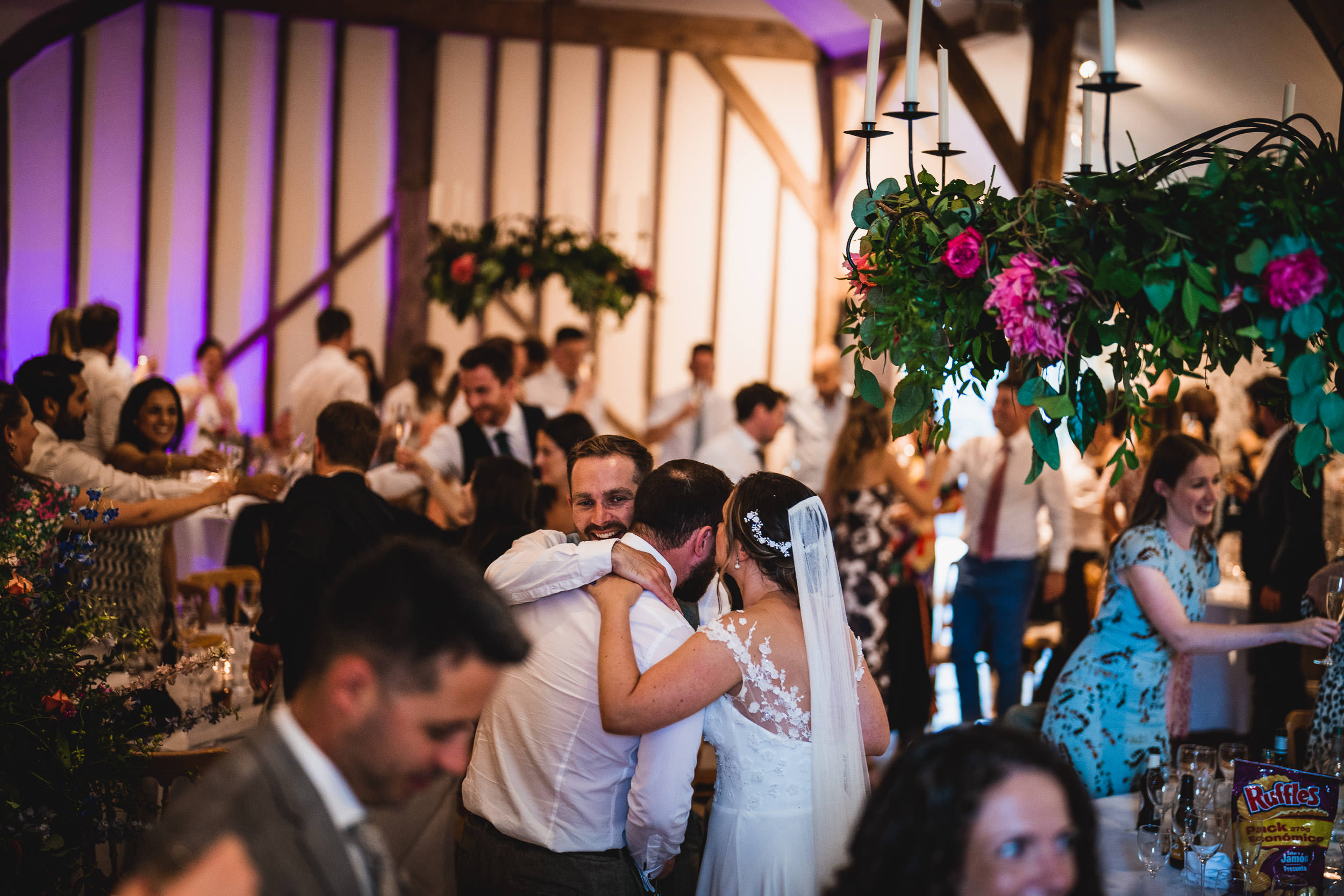 A wedding reception with guests socializing. A couple embraces in the foreground, surrounded by floral decorations and chandeliers.