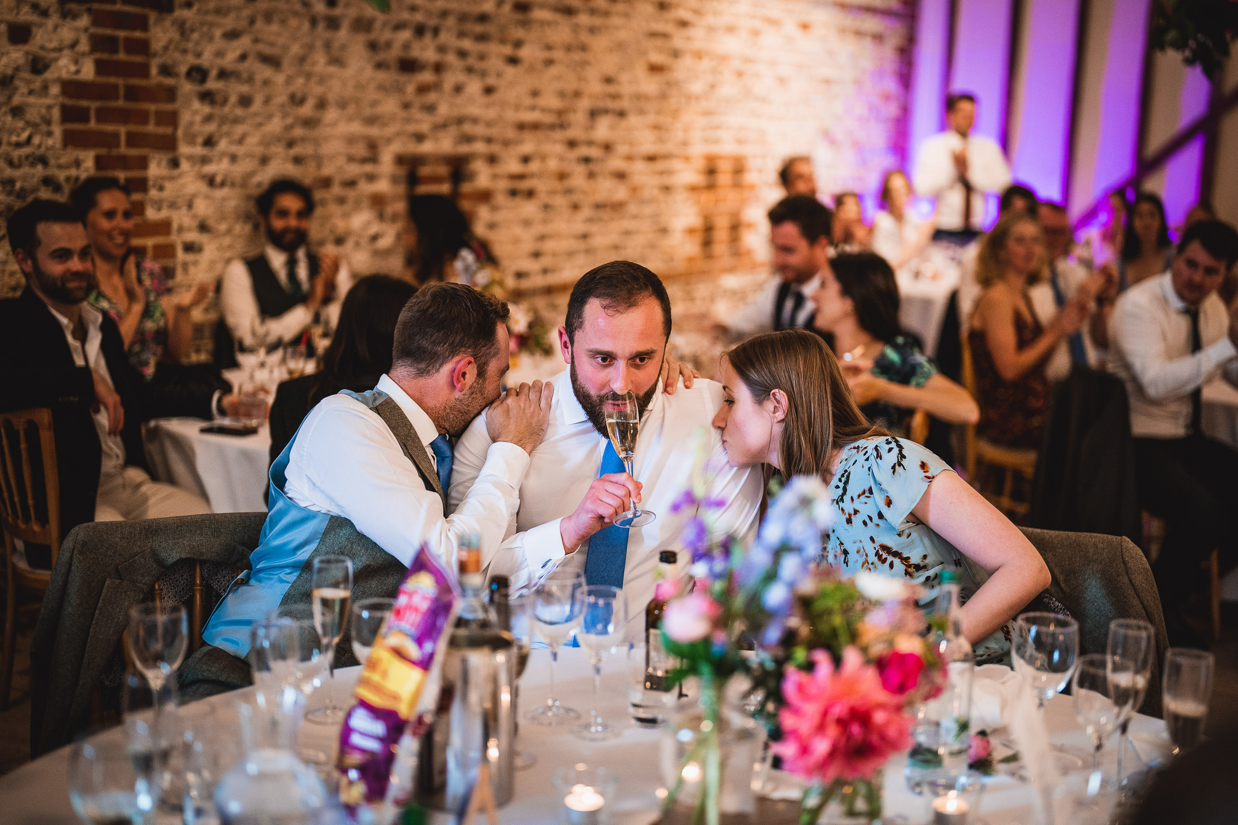 People seated at a dining table in a dimly lit event space, sharing a drink and conversation. The table is decorated with flowers and glasses.