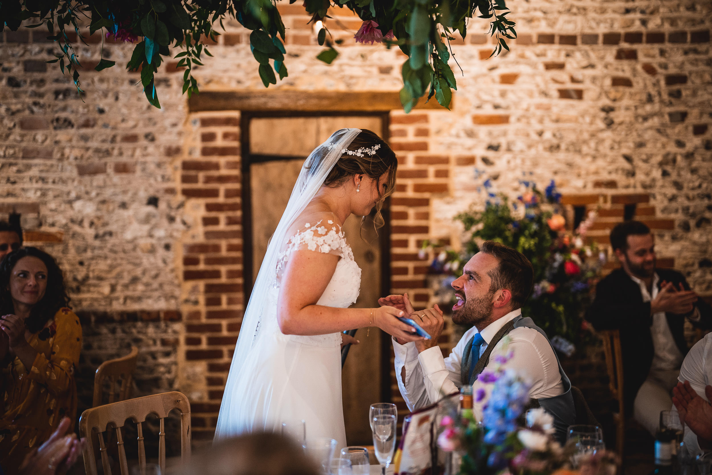 A bride and groom share a moment during their wedding reception, surrounded by guests and floral decorations in a rustic setting.