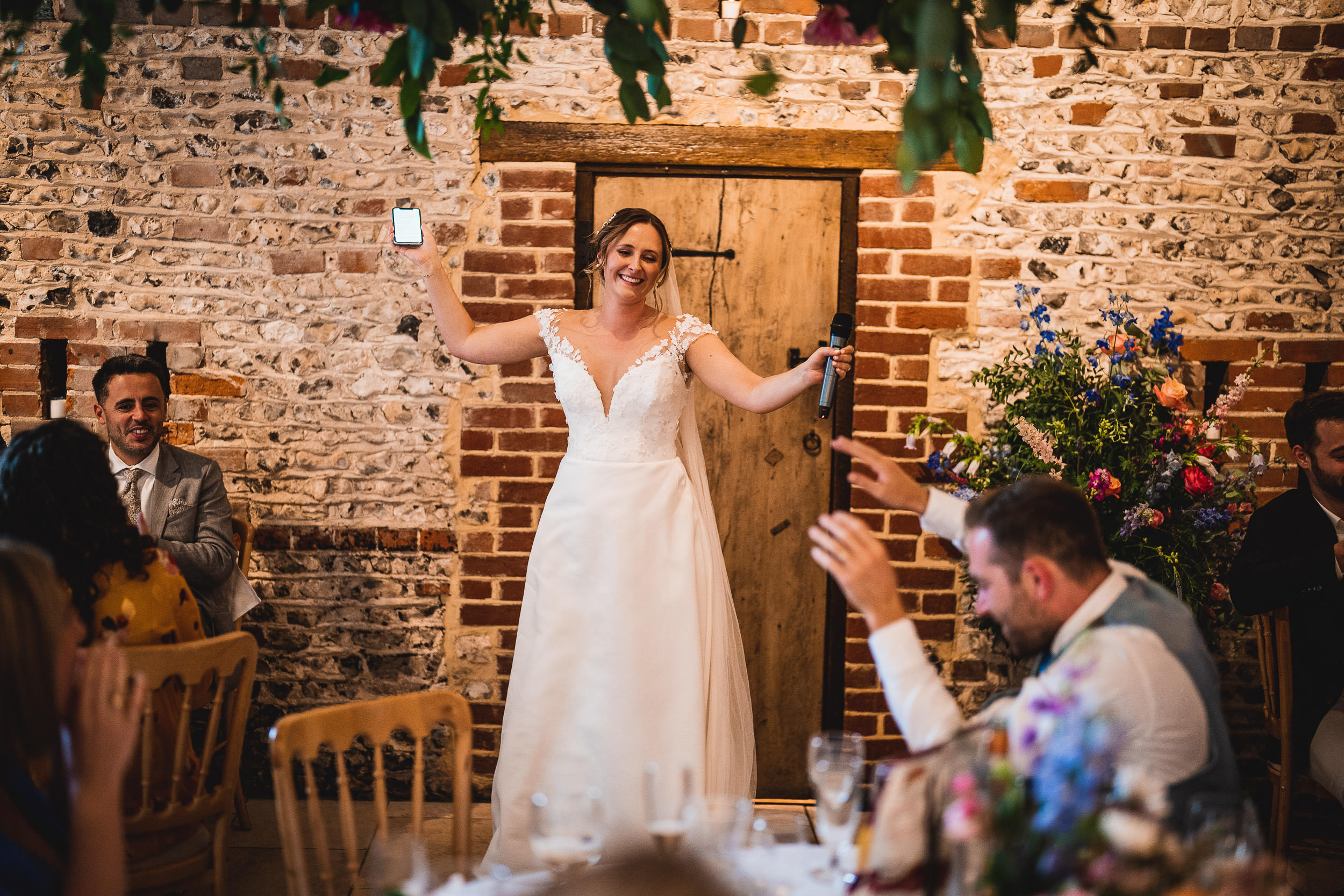 A bride in a white gown holds a microphone and a phone, smiling. Guests sit and gesture toward her in a rustic venue with brick walls and floral decorations.