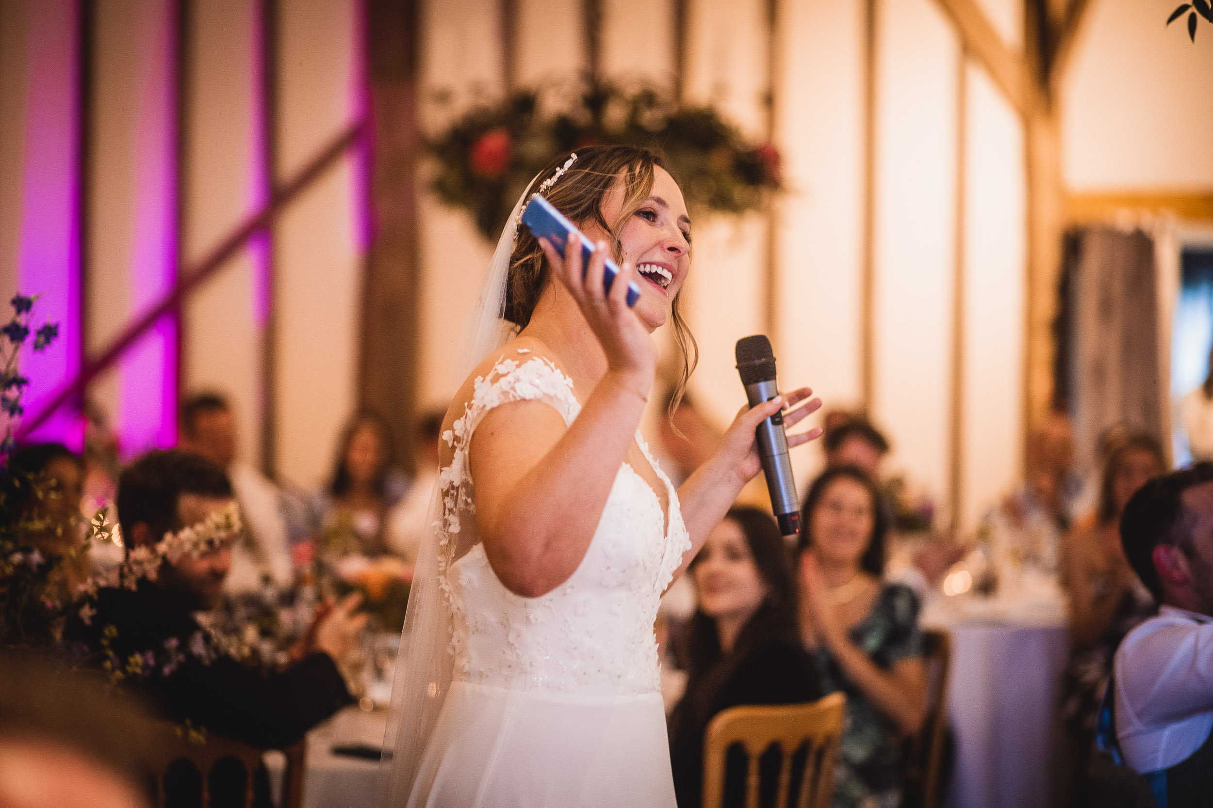 Bride in a white wedding dress holds a microphone and phone, speaking and smiling. Guests seated at tables in the background.