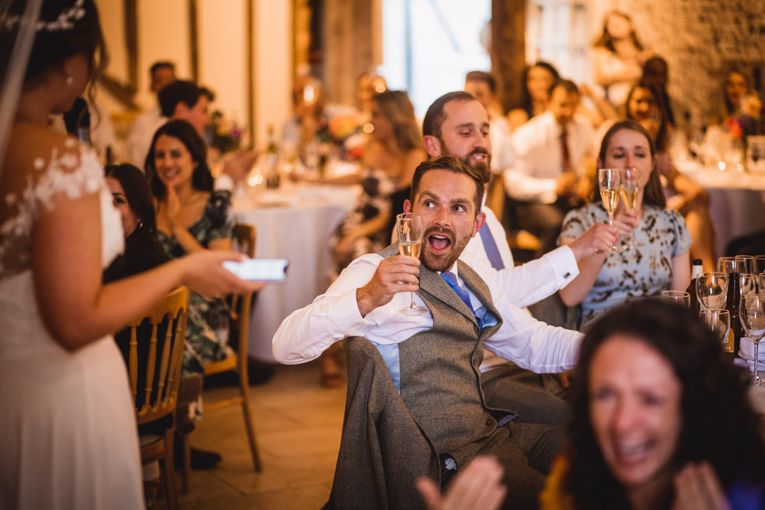 A man in a suit excitedly holds up a champagne glass while seated among guests at a wedding reception.