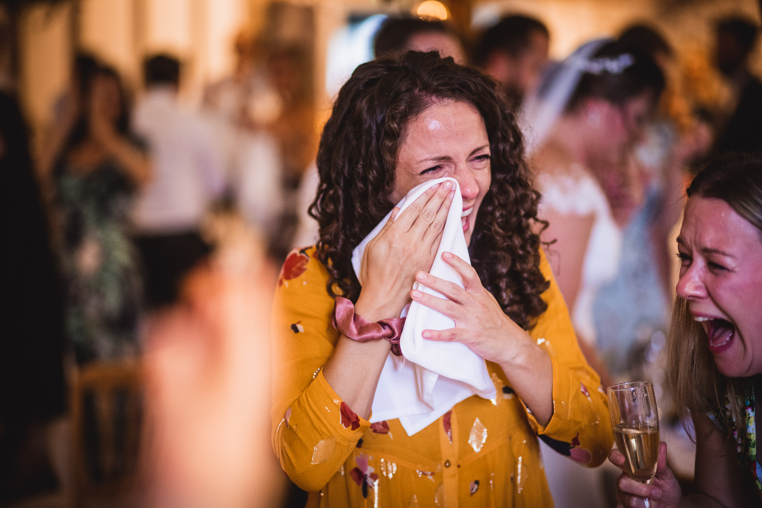 A woman in a yellow dress is laughing and wiping her tears with a napkin at a crowded celebratory event.