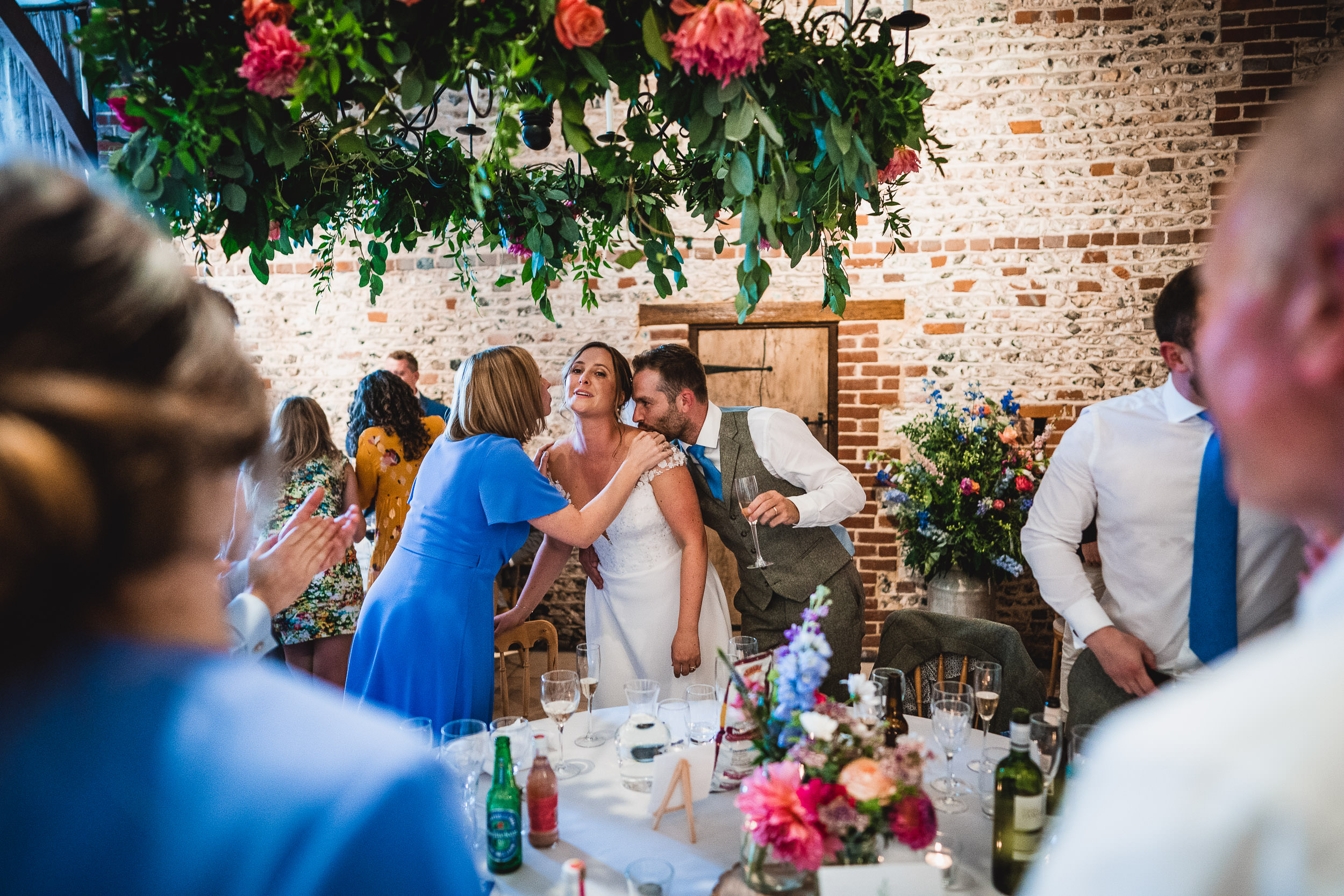 Bride in a white dress is congratulated by a woman in blue, while a man in a vest smiles beside her. The setting is a rustic venue with floral decorations and guests around a table.