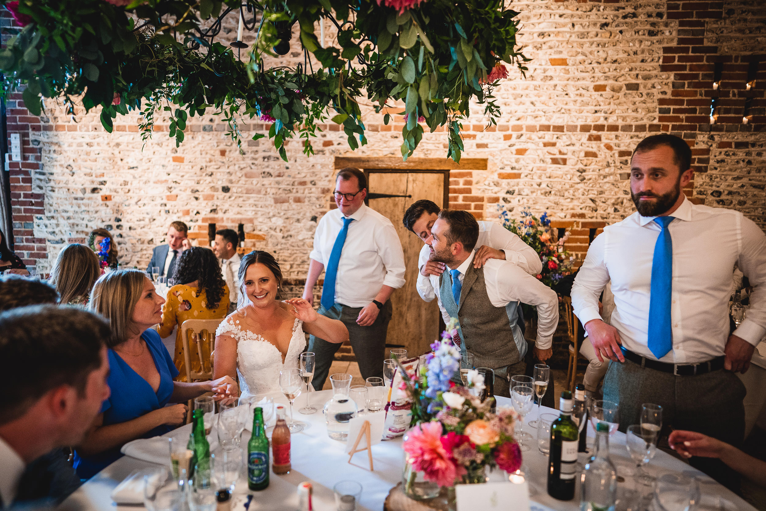 A bride sits at a decorated table with guests. People are laughing and talking. There are drinks and flowers on the table, and greenery hangs above.