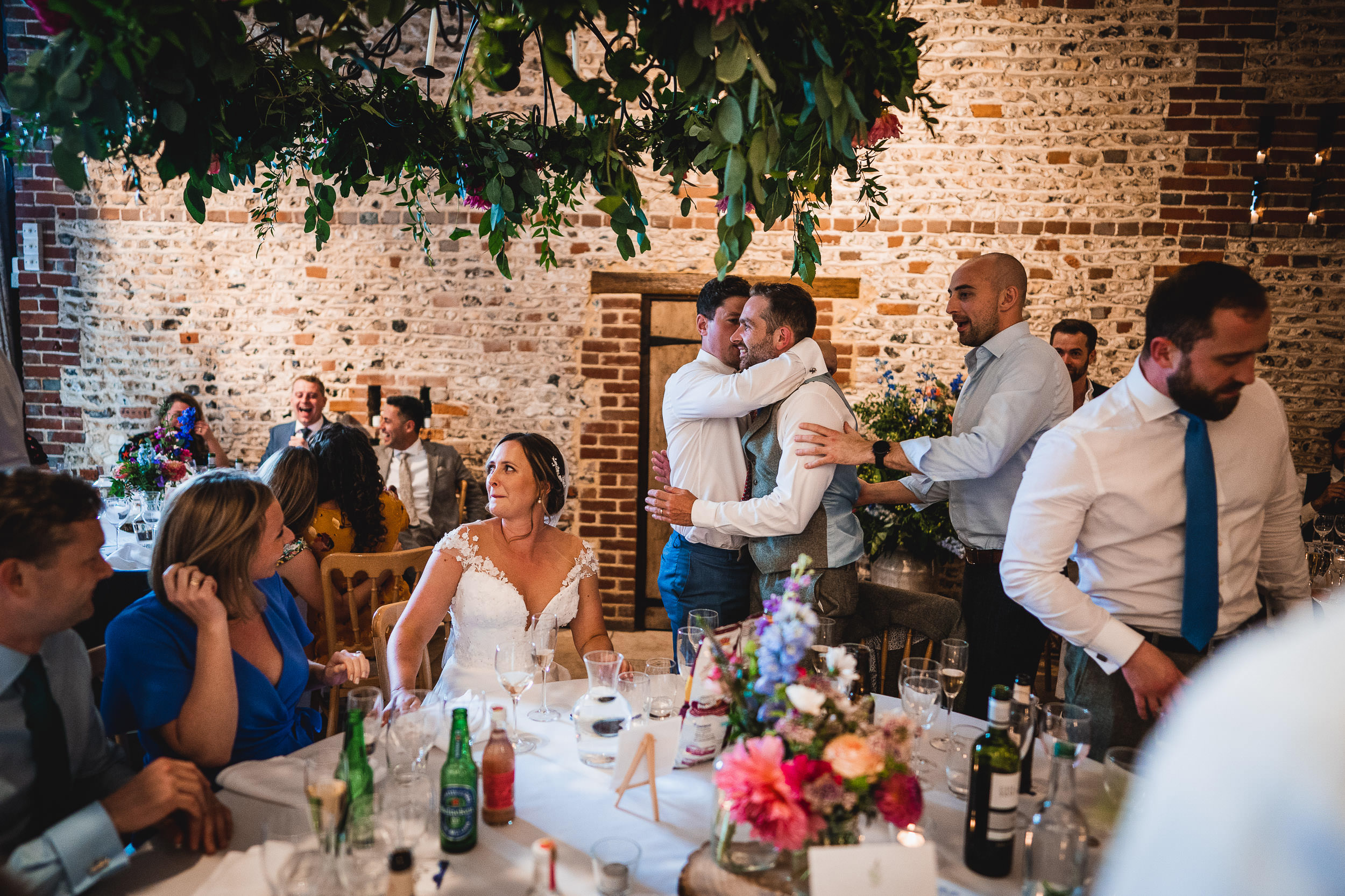 Guests converse and embrace at a wedding reception with a brick wall and floral decor in the background.