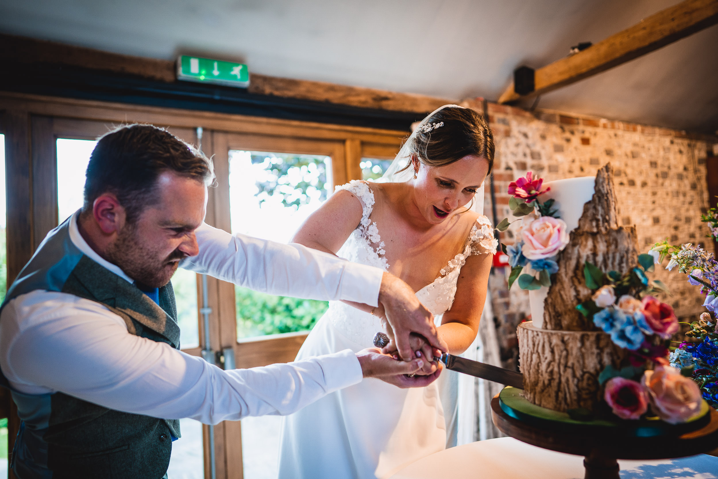 A bride and groom are cutting a wedding cake decorated with flowers and a tree stump design, indoors next to a large window.