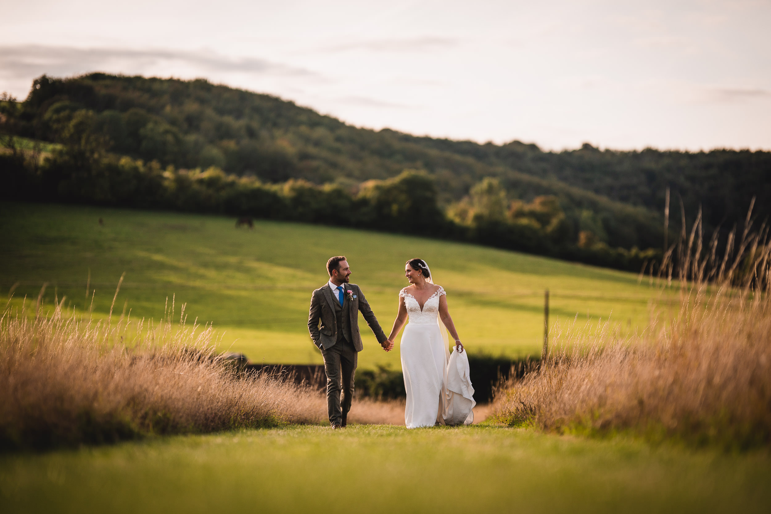 A couple in wedding attire walks hand in hand through a grassy field with hills in the background.