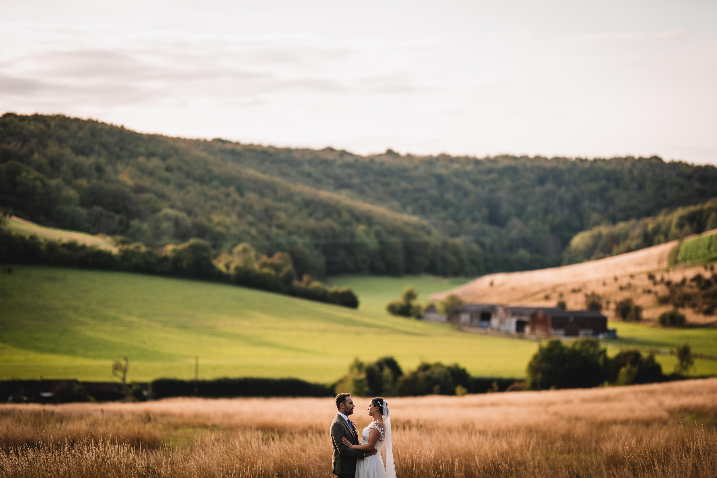 A couple stands together in a grassy field with rolling hills and trees in the background. The sky is partly cloudy.