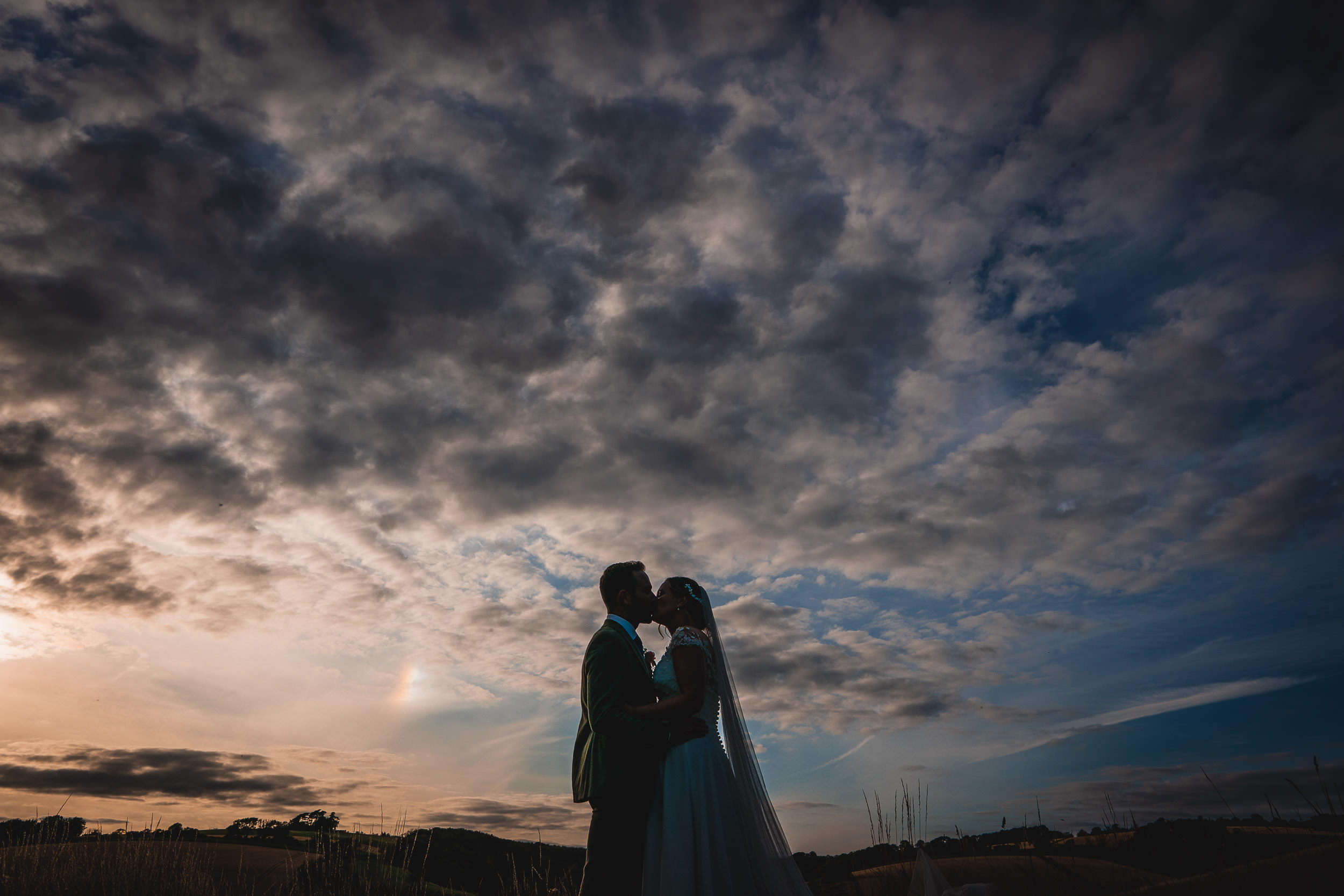 Silhouetted couple kissing at sunset under a dramatic, cloudy sky.