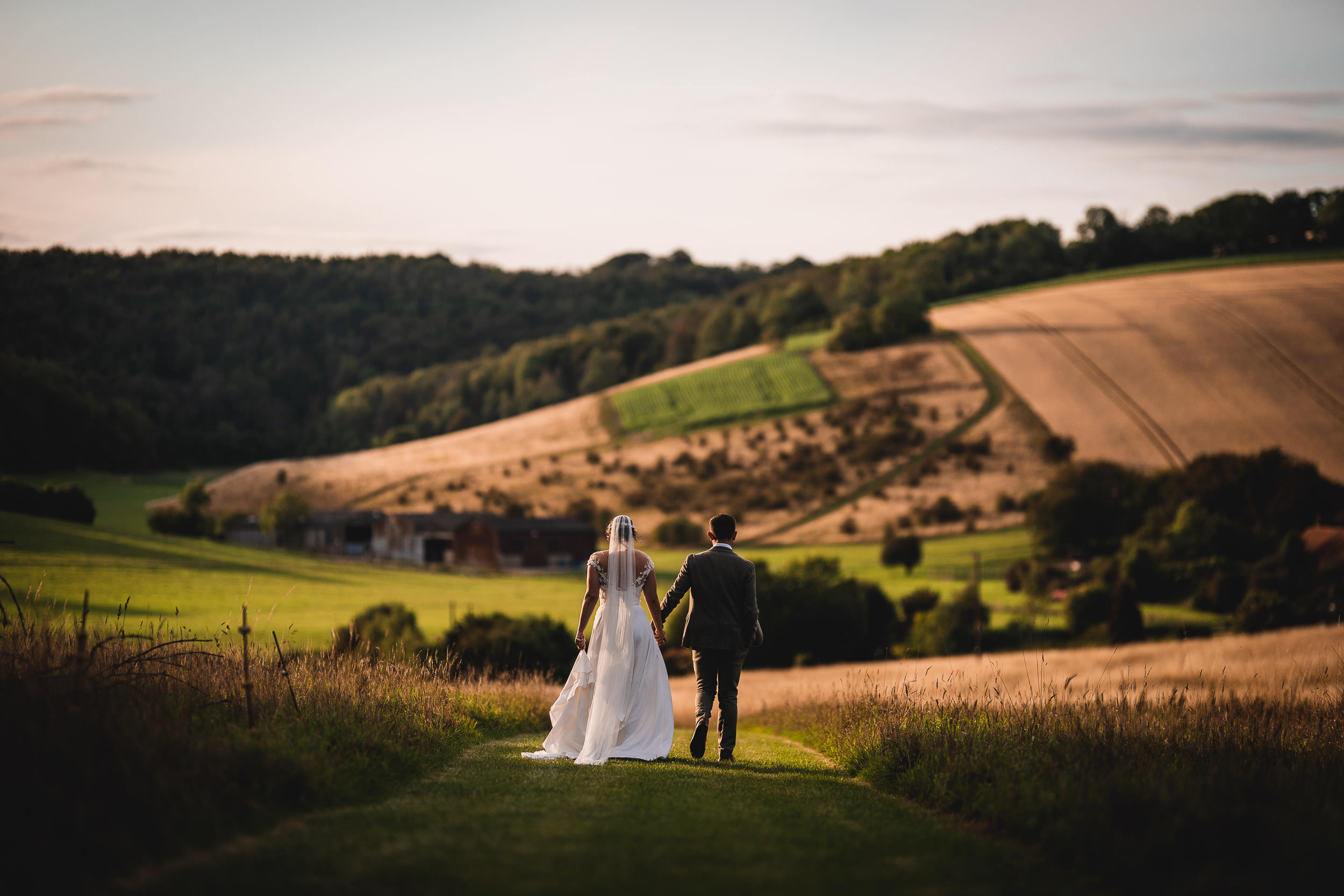 A bride and groom walk hand in hand down a grassy path with rolling hills and farm fields in the background.