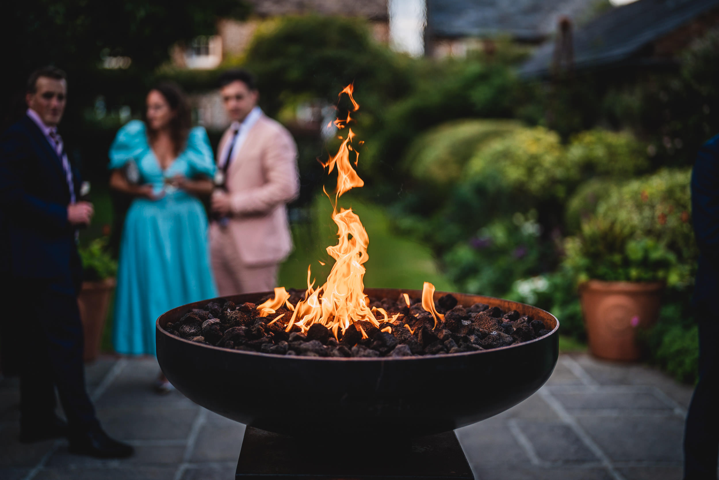 A fire pit with flames burning in the foreground, with people in formal attire chatting in the blurred background.