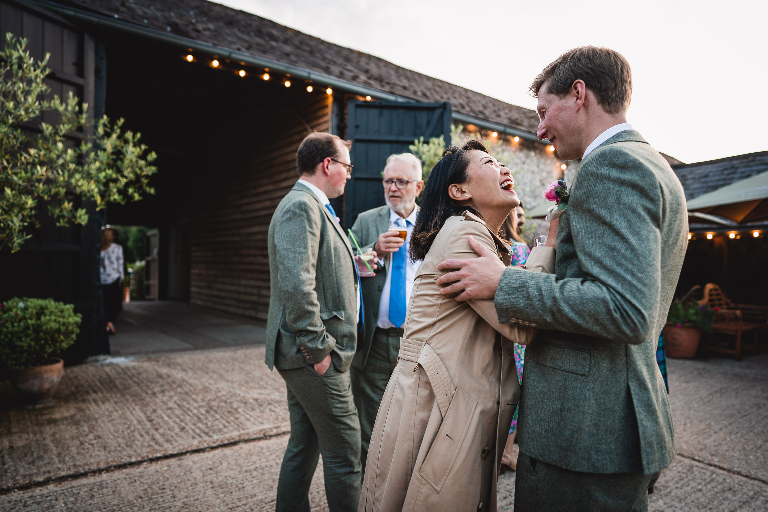 A group of people in formal attire are laughing and talking outside a rustic building with string lights.