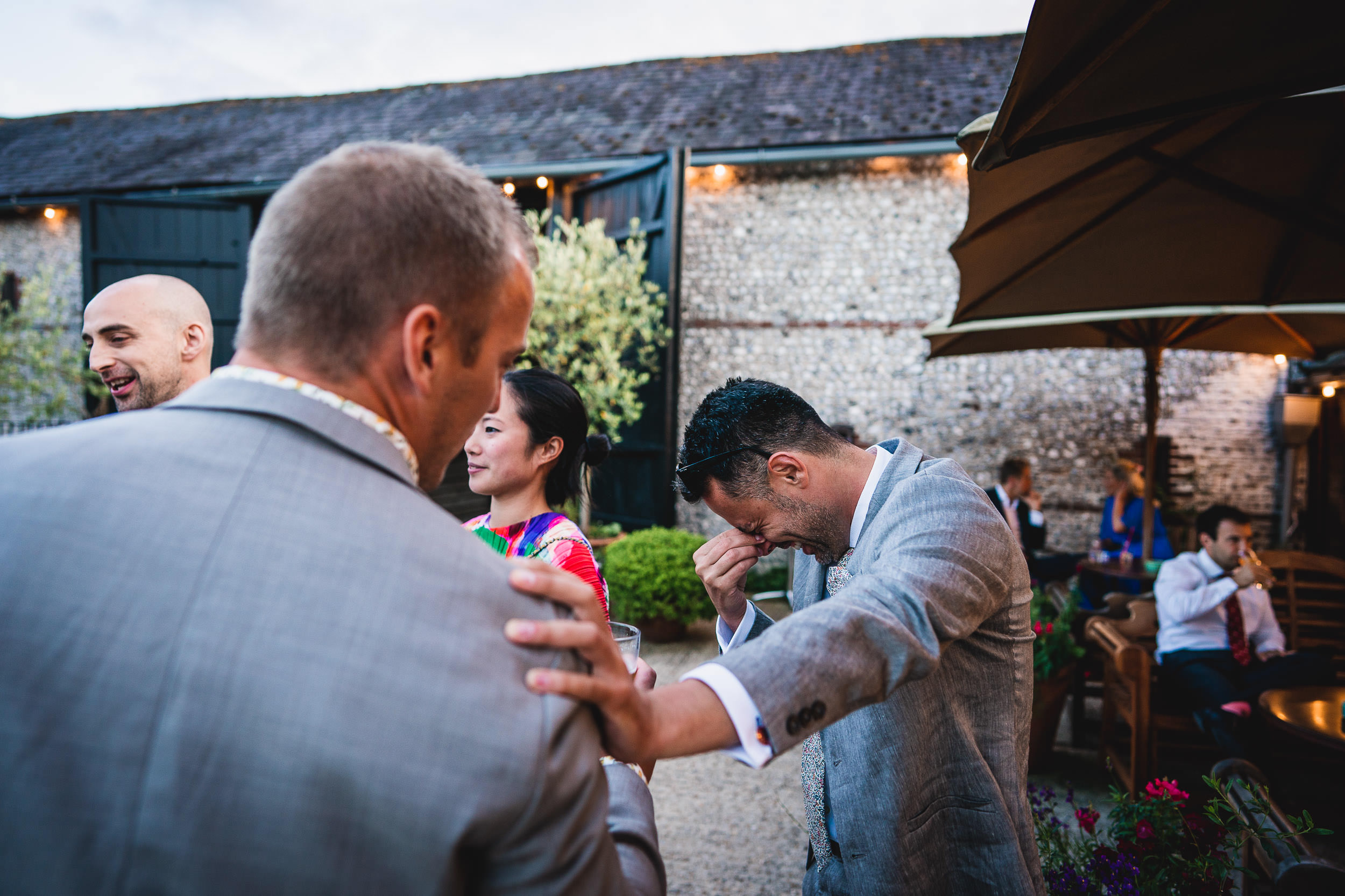 A group of people in formal attire are gathered outside. One man is holding his face, appearing emotional, while another man pats his shoulder. The surrounding area has greenery and patio seating.