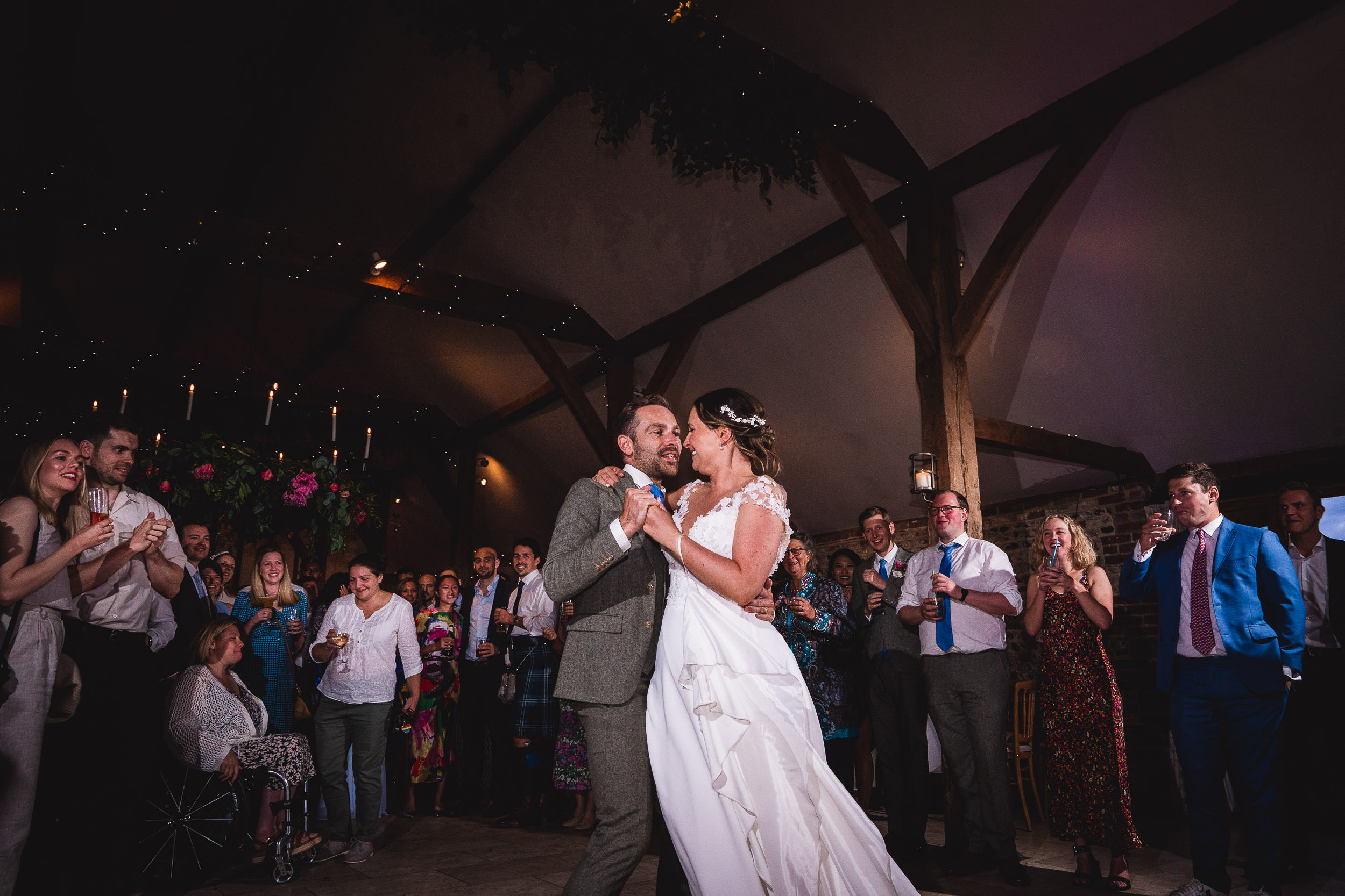 A bride and groom dance in a rustic venue surrounded by guests, some clapping and smiling.