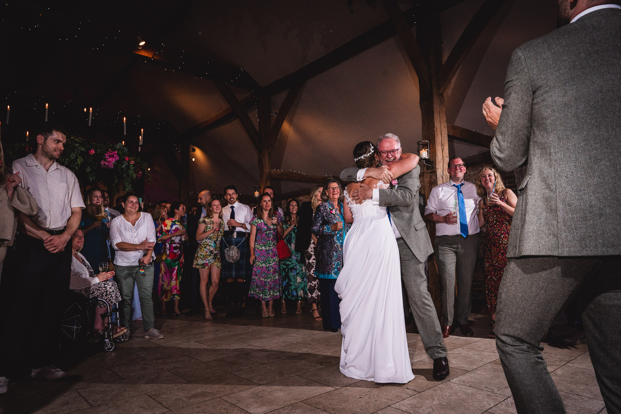 A bride and an older man embrace on a dance floor, surrounded by smiling guests in colorful attire at a wedding reception.