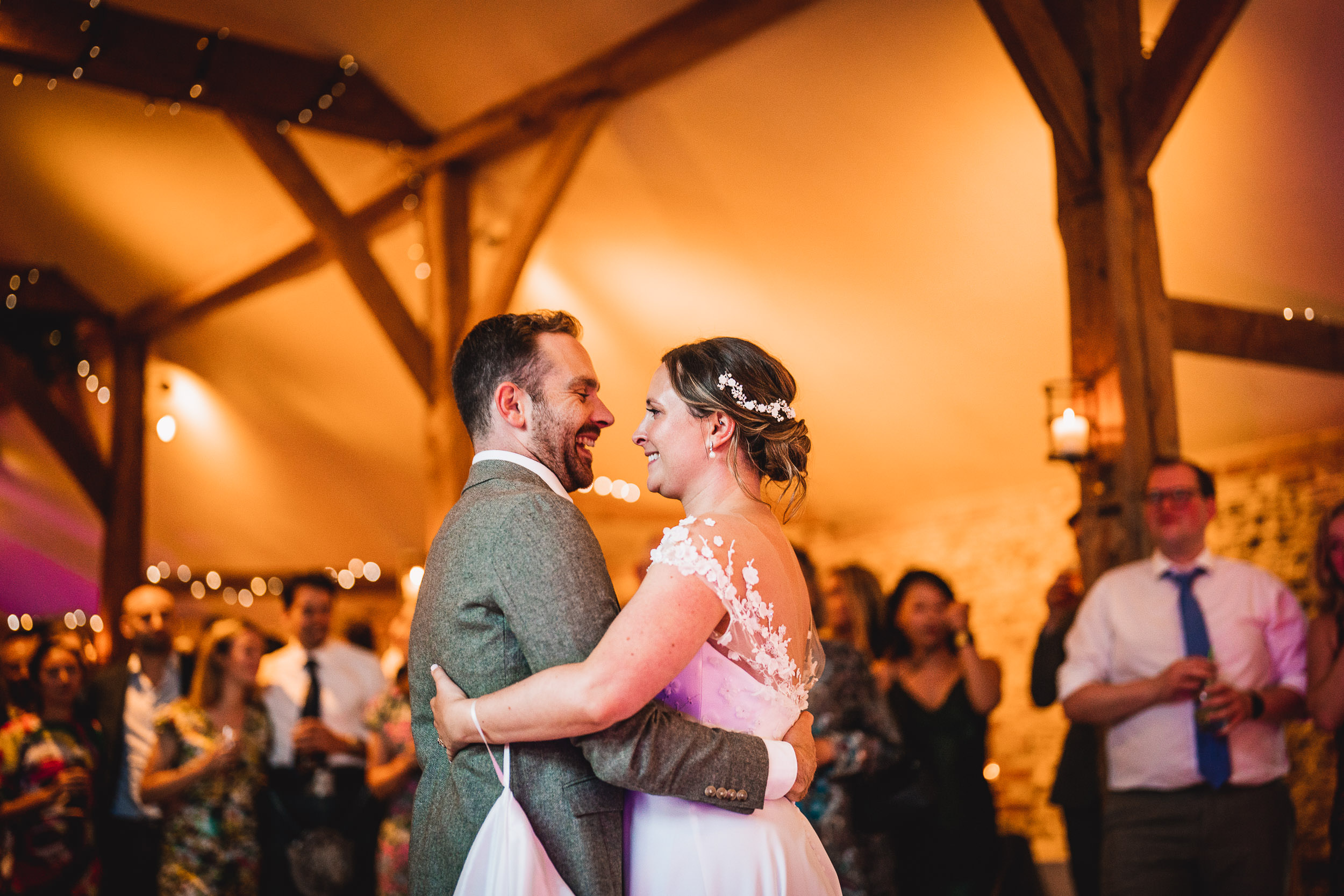 A bride and groom share their first dance in a warmly lit venue, surrounded by smiling guests.